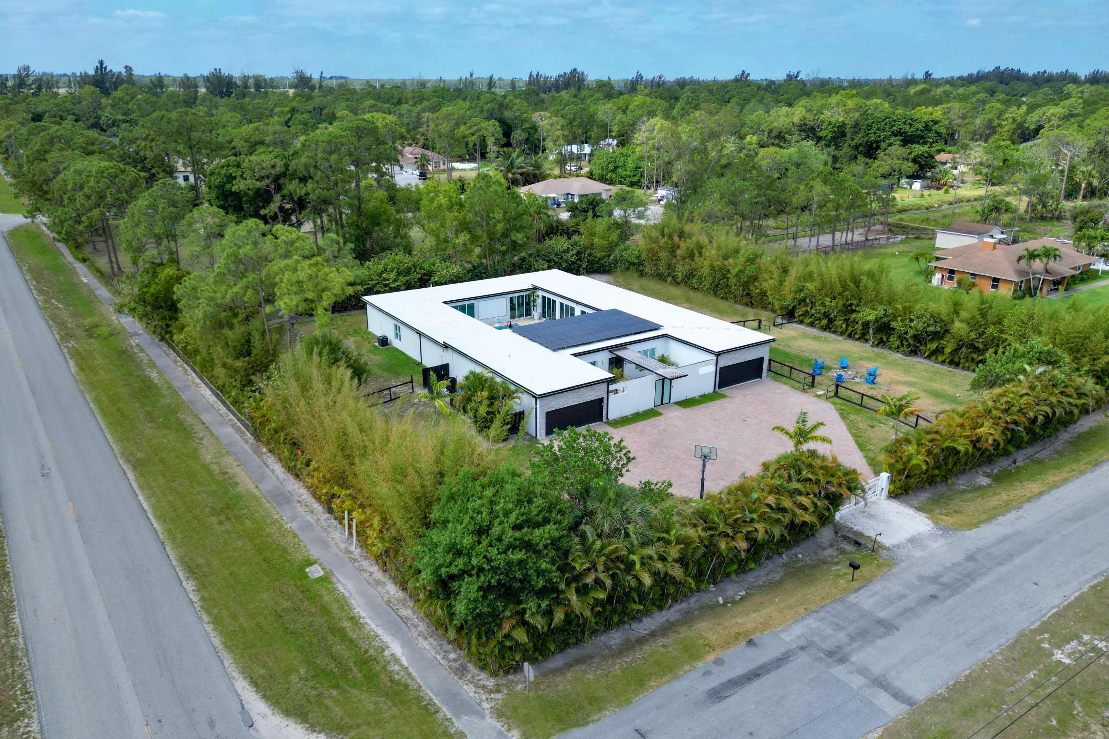 The image shows a modern, single-story house surrounded by greenery, situated at the intersection of two roads.