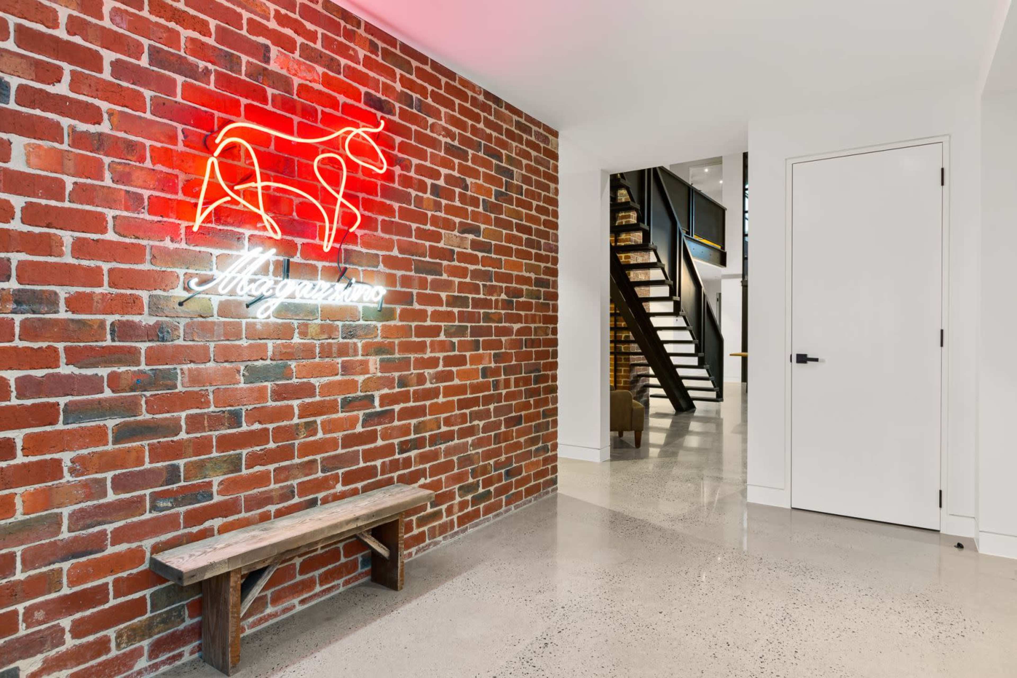 A hallway with a brick wall featuring a neon horse sign, a wooden bench, and a staircase in the background.