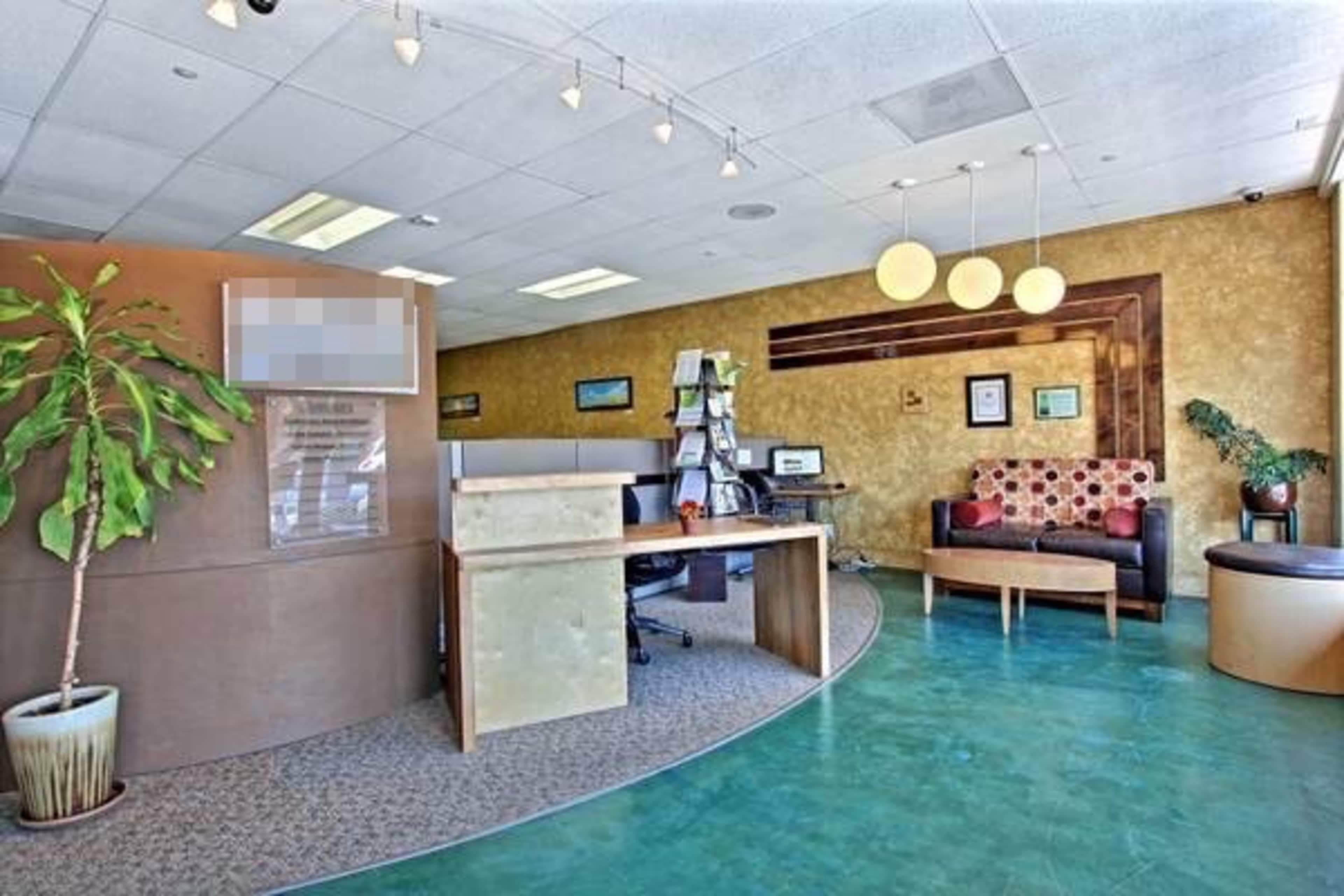 An office reception area with a circular layout, featuring a front desk, a seating area with a patterned couch, and a potted plant.