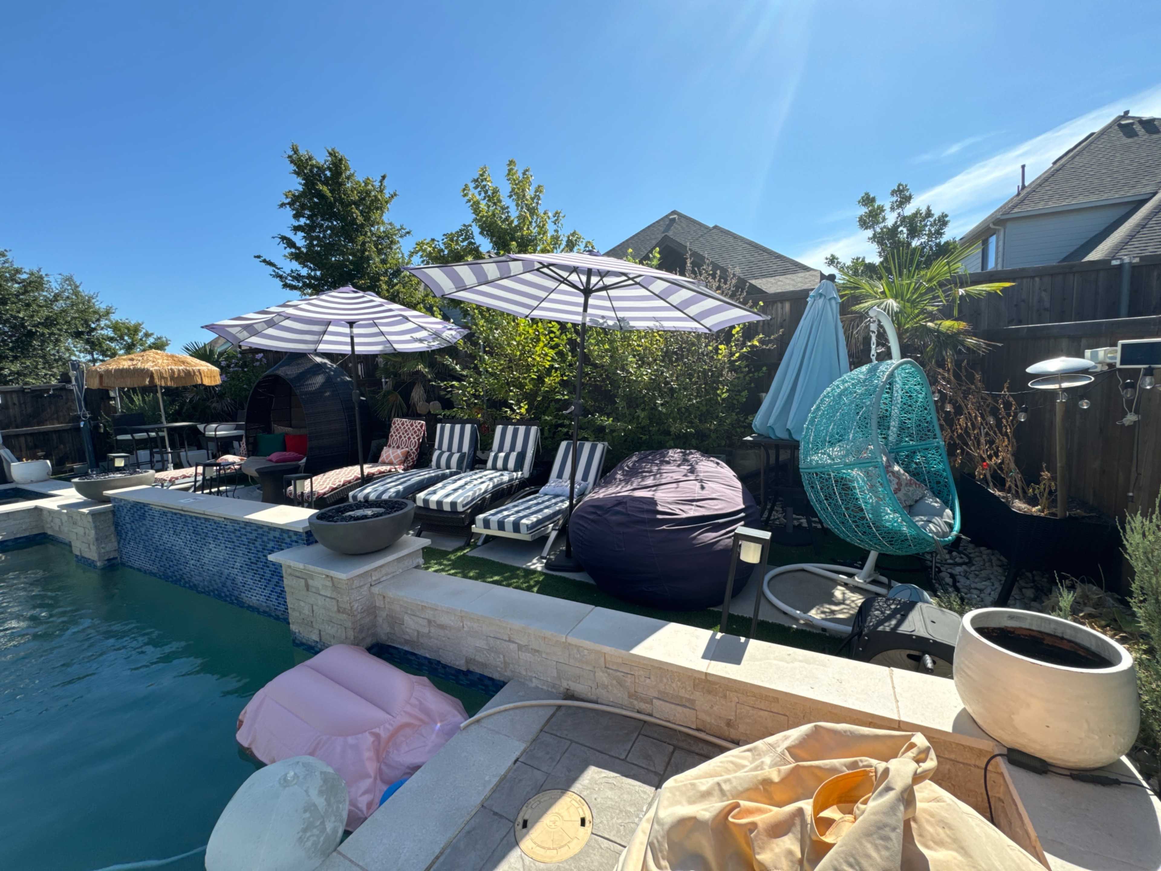 The image shows a backyard pool area with striped lounge chairs, large umbrellas, a hanging chair, and a variety of greenery.