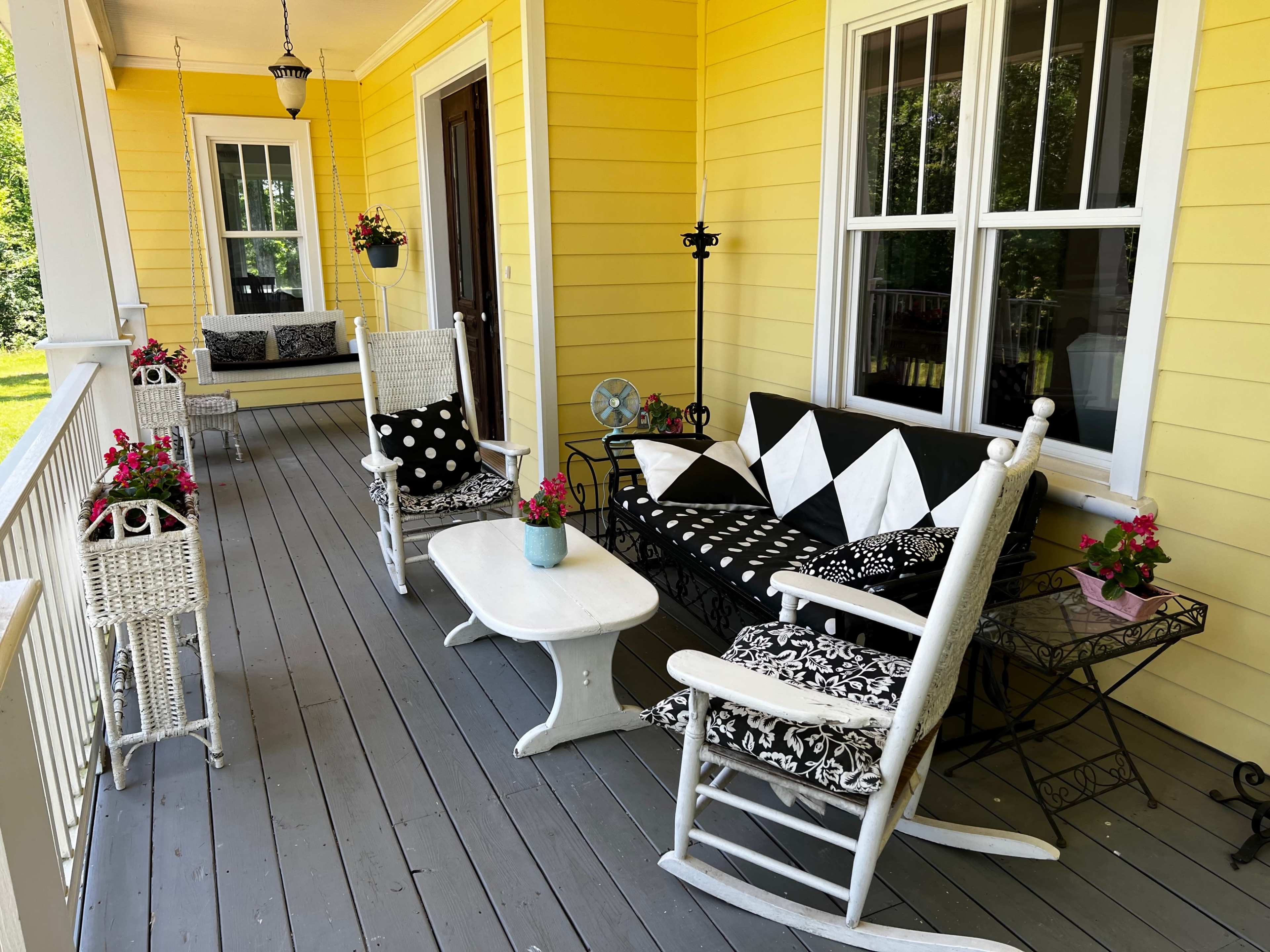 A bright yellow porch featuring white rocking chairs, a black and white upholstered bench, a small white table, and various potted flowers.