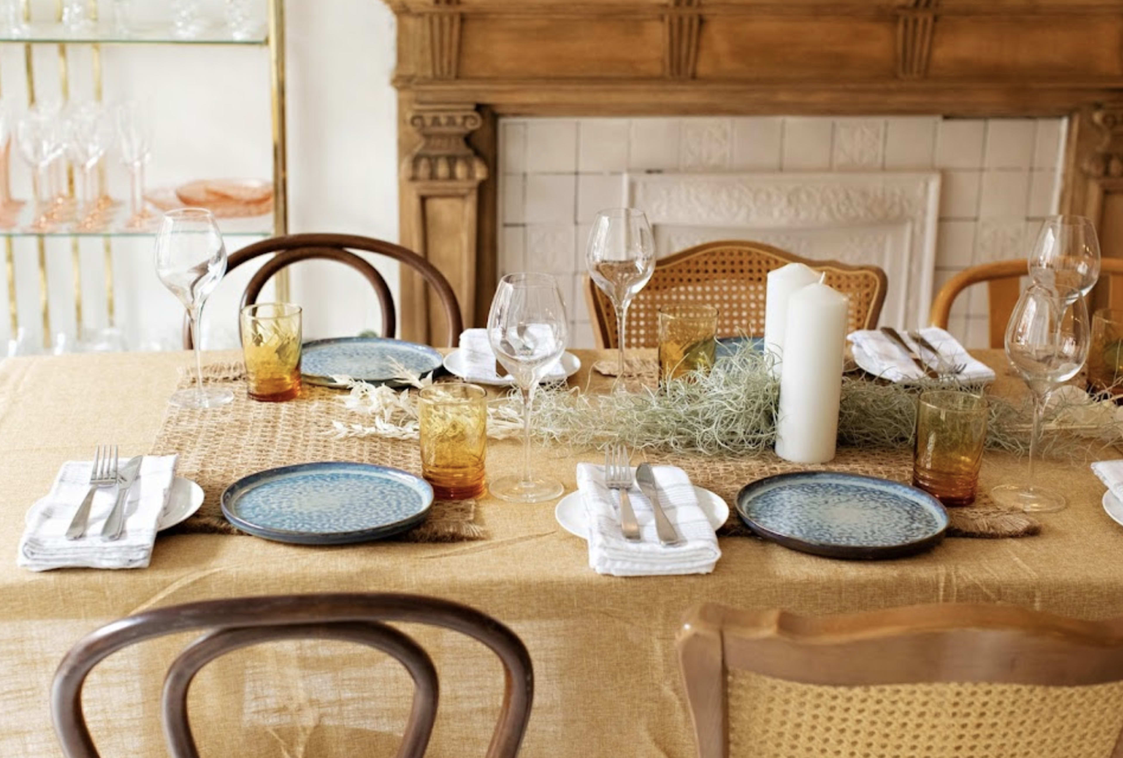 A dining table is set with blue patterned plates, glassware, and candles, decorated with a woven runner and greenery.