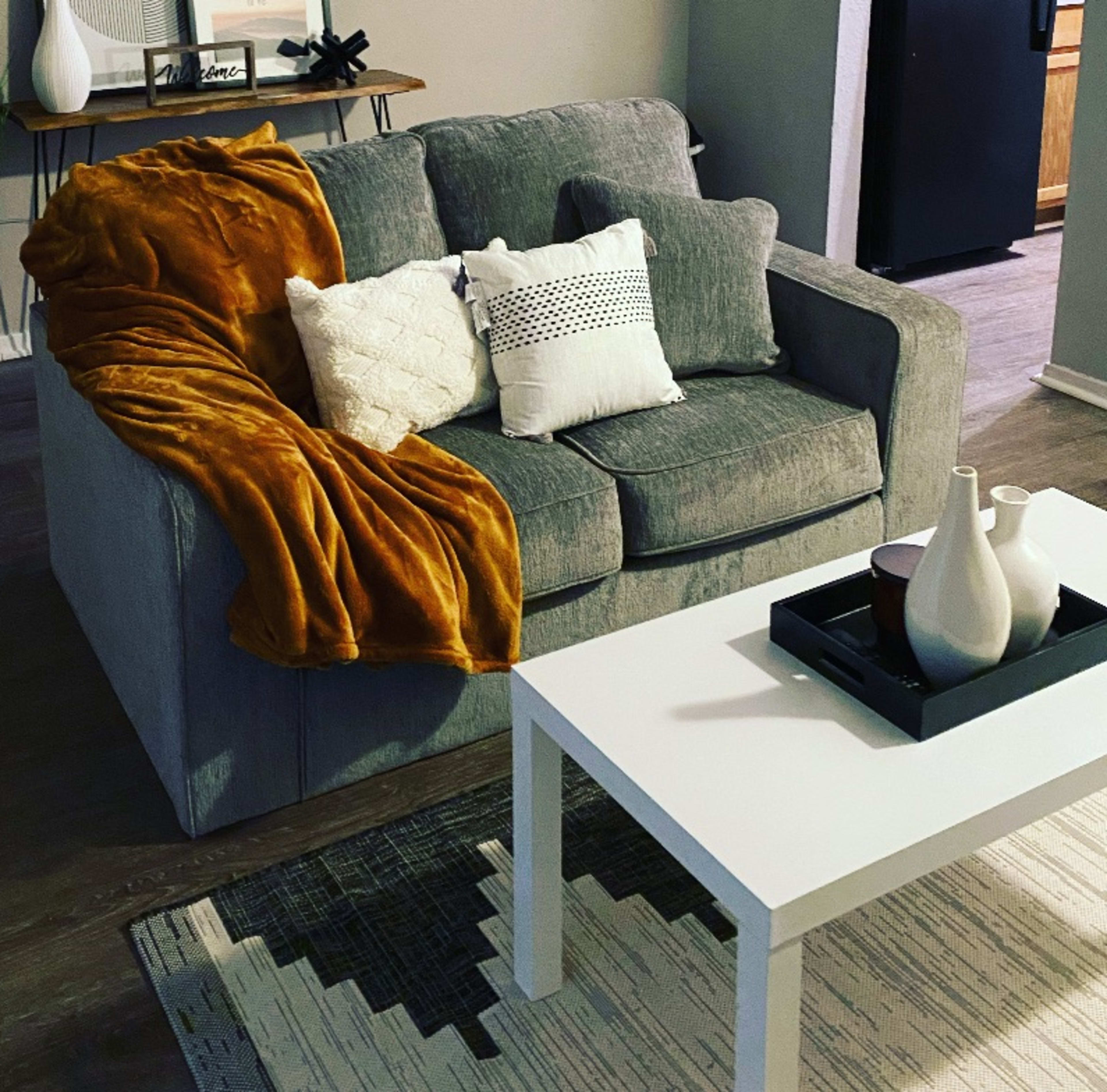 A gray couch with a mustard-colored throw blanket and several decorative pillows is positioned beside a white coffee table on a patterned rug in a living room.