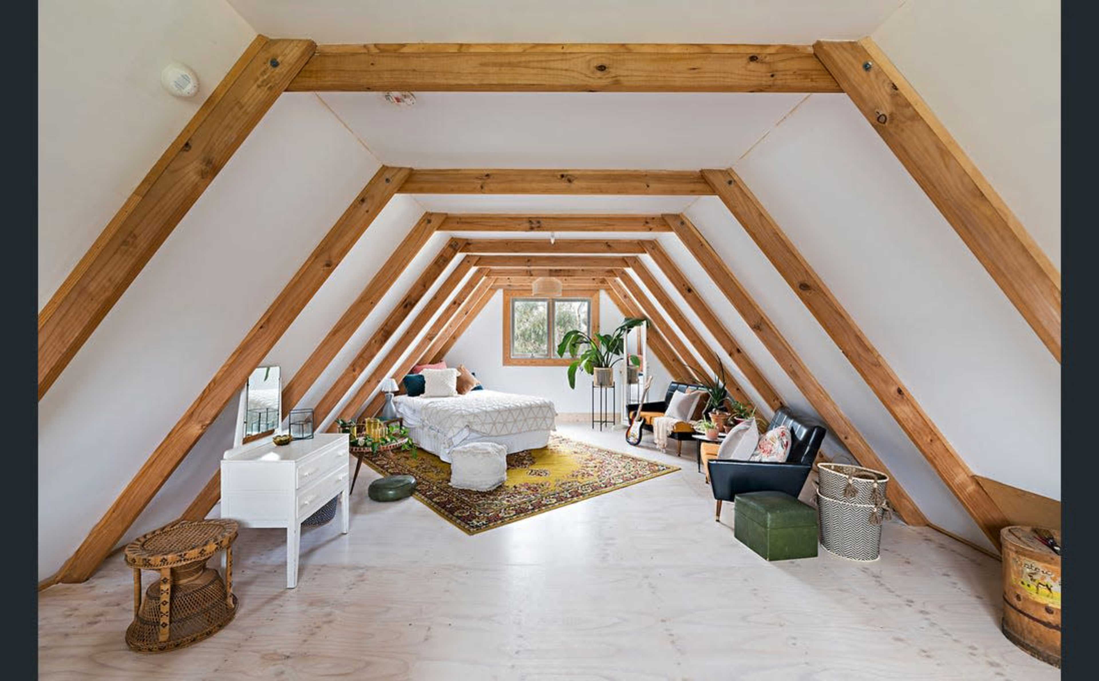 A spacious attic room with wooden beams, featuring a bed, a small sofa, a side table, and plants near a window.