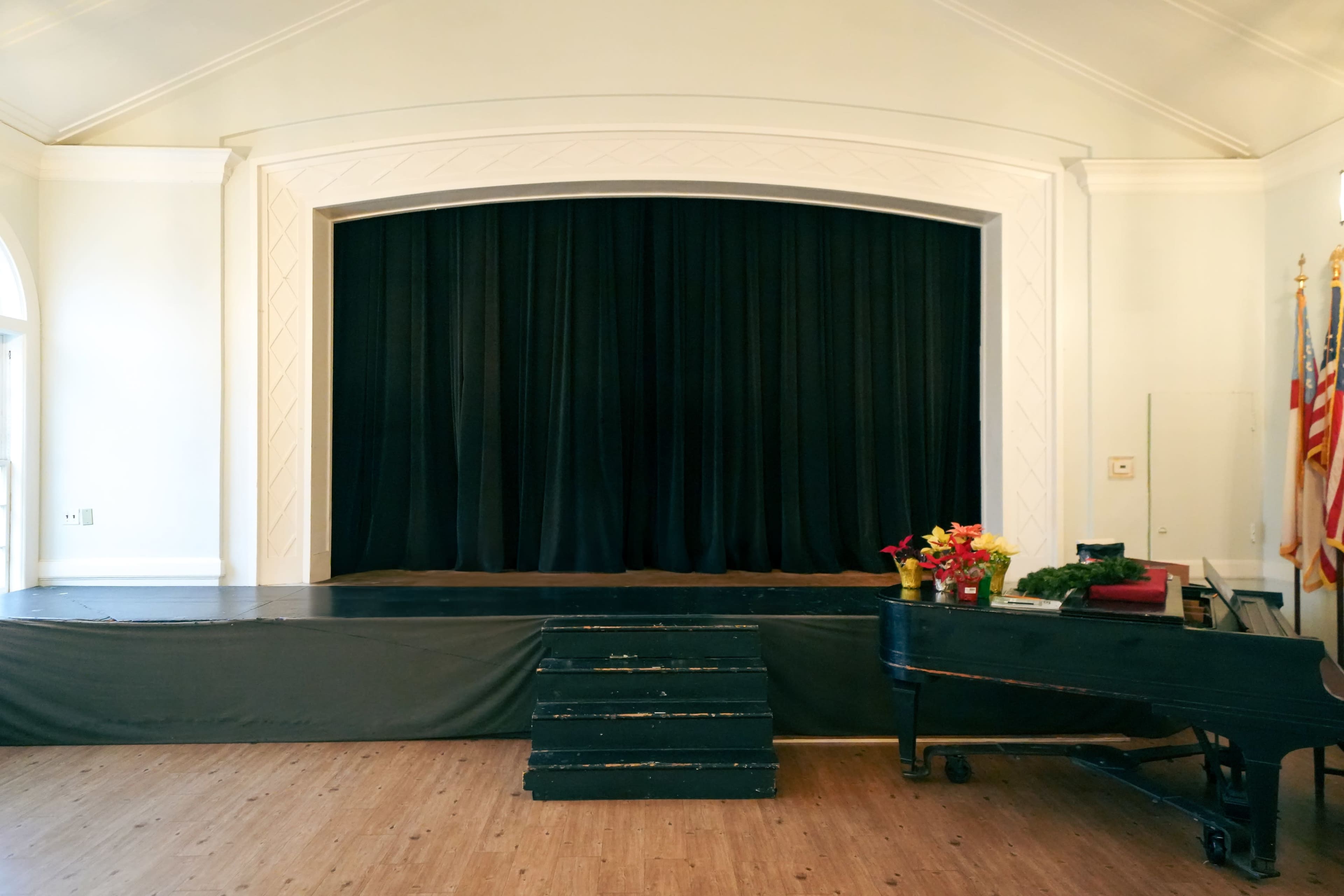 The image shows a stage with a black curtain, flanked by flags, and a piano positioned to the right.