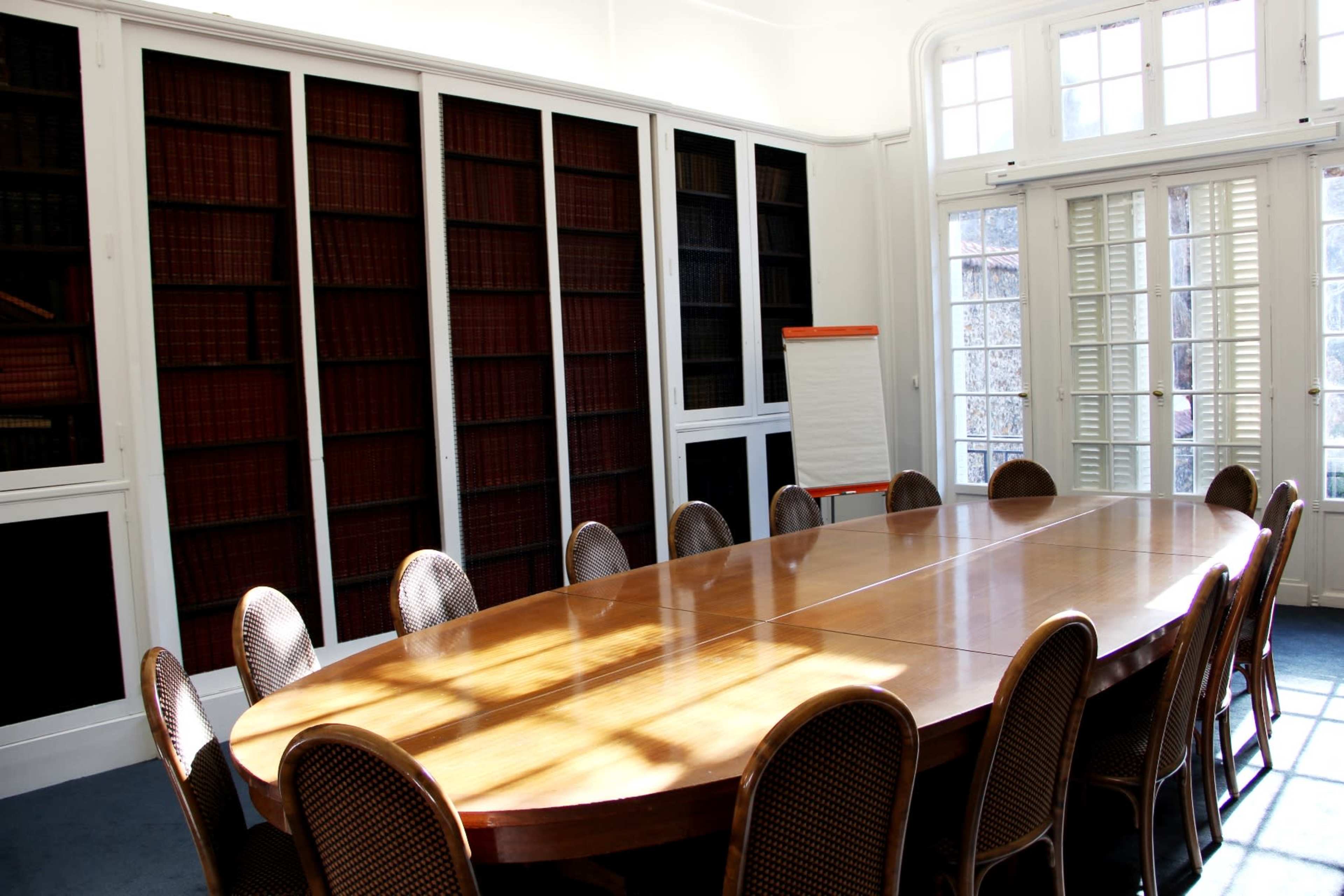 A large oval wooden table surrounded by chairs is situated in a well-lit room with bookshelves lined with red volumes.