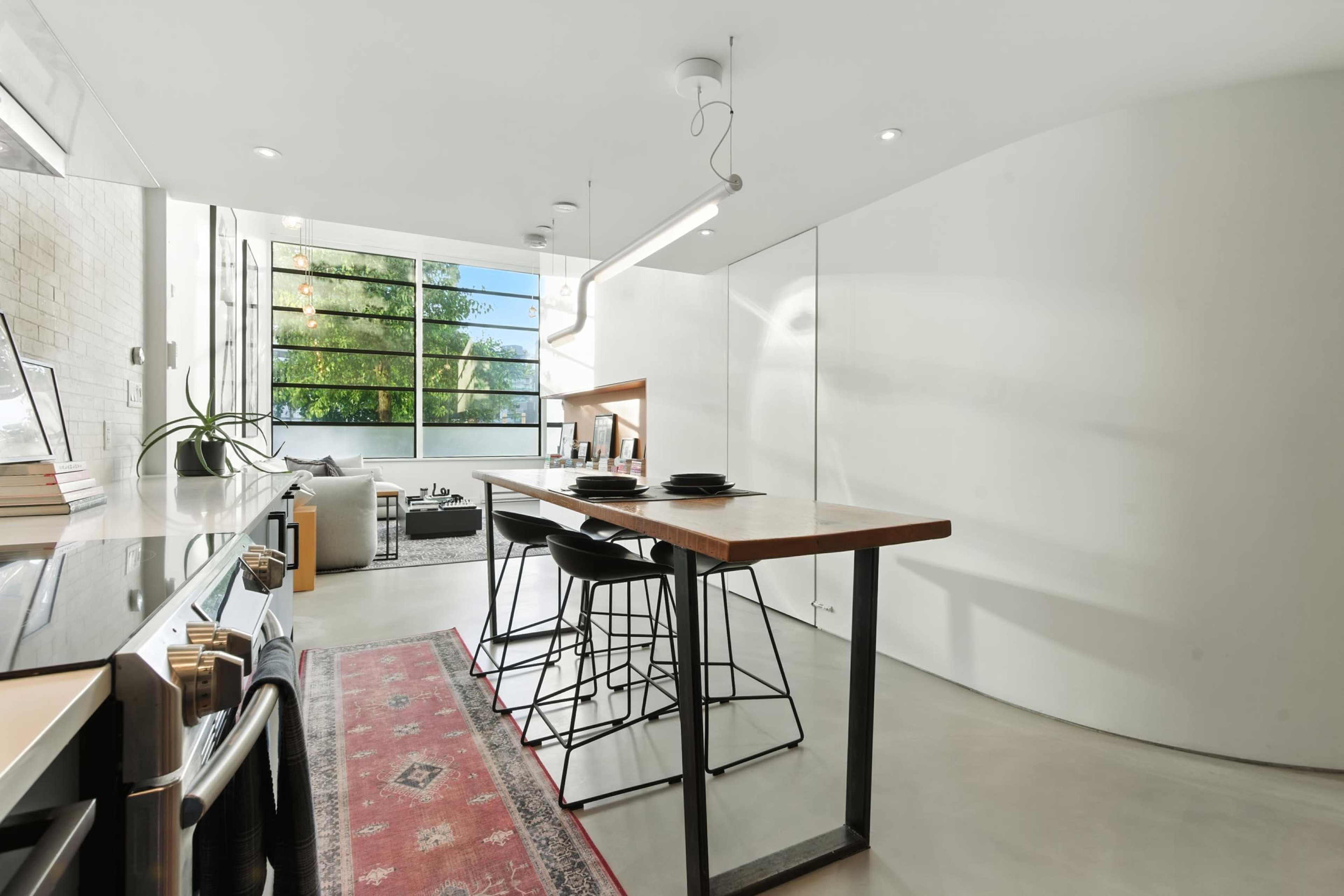 The image shows a modern kitchen and dining area with a long table, black bar stools, and large windows allowing natural light to fill the space.