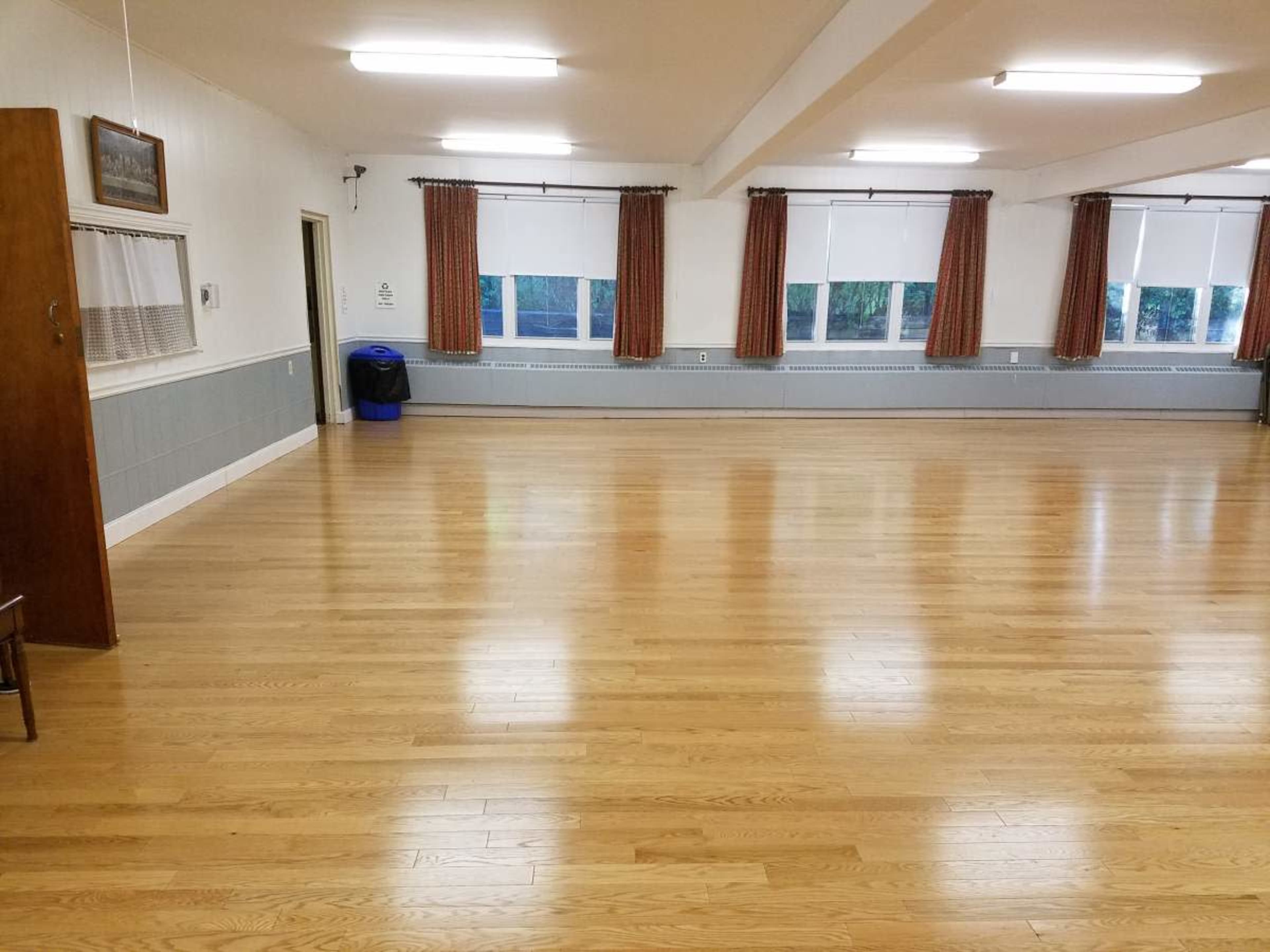 The image shows a spacious, empty room with polished wooden floors, large windows, and curtains.