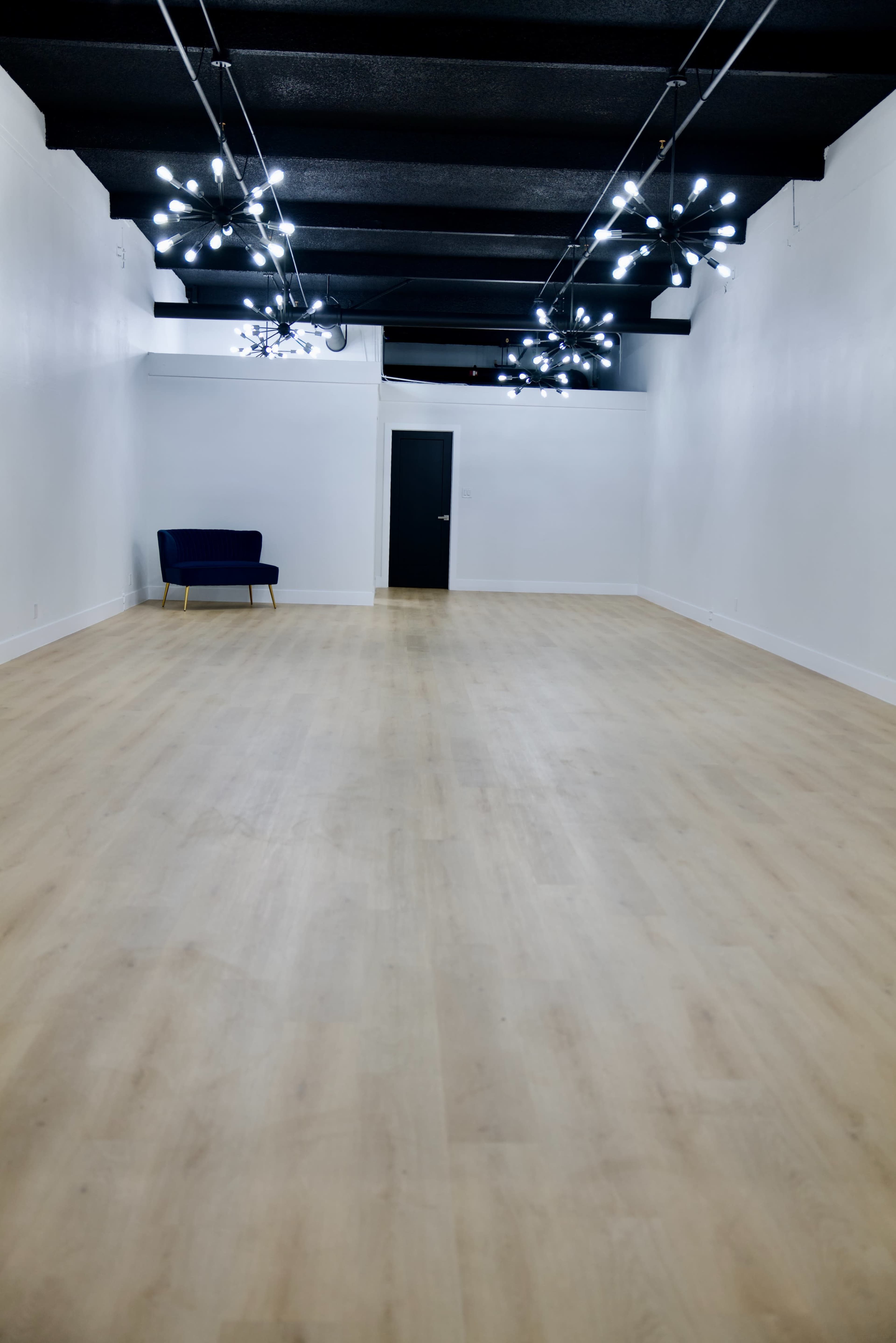 The image shows a spacious, empty room with light wooden flooring, white walls, and modern pendant lights, featuring a single dark blue chair in the corner.