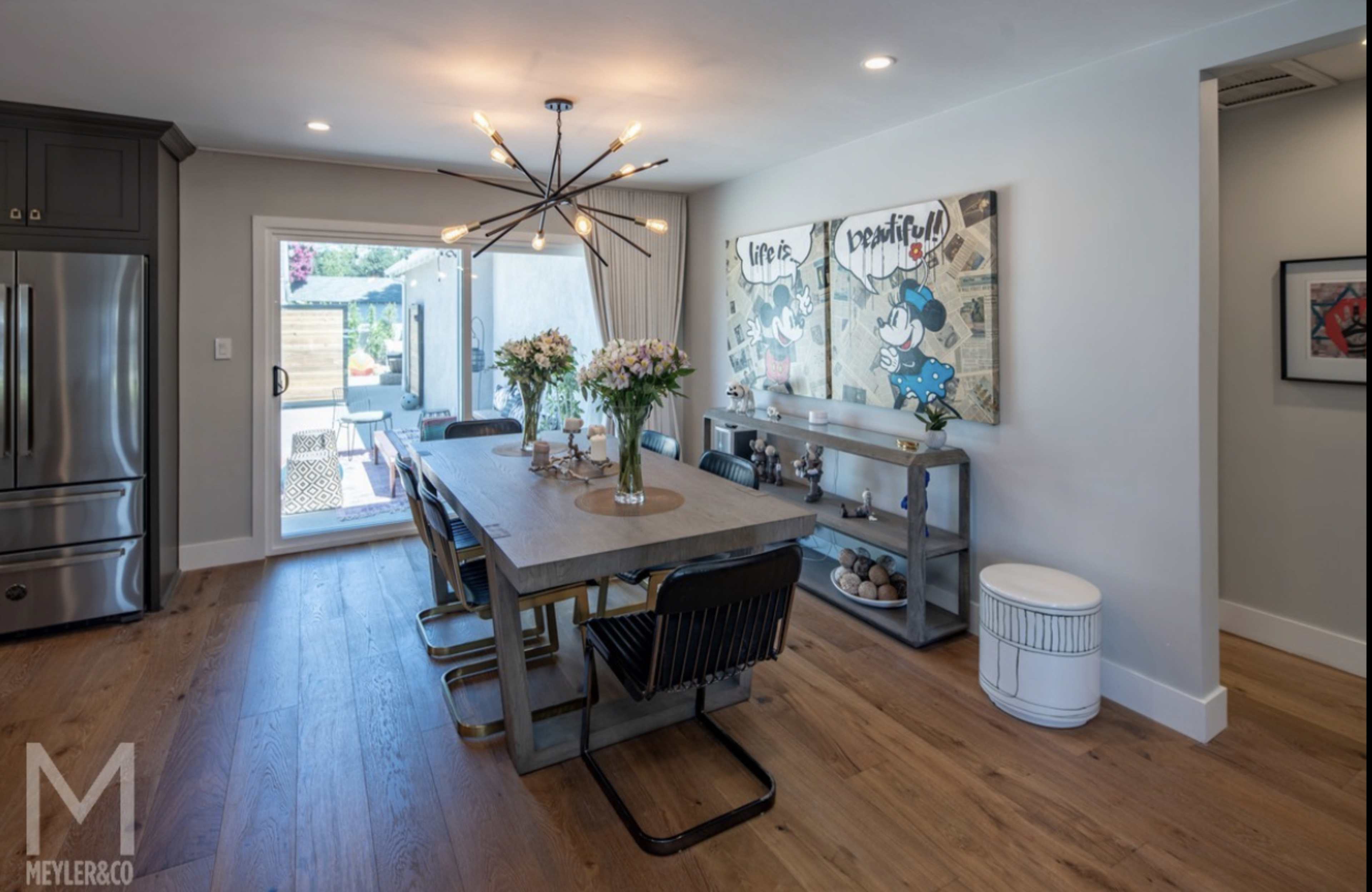 A modern dining area with a long table, black chairs, and two large artwork pieces on the wall, illuminated by an overhead light fixture.