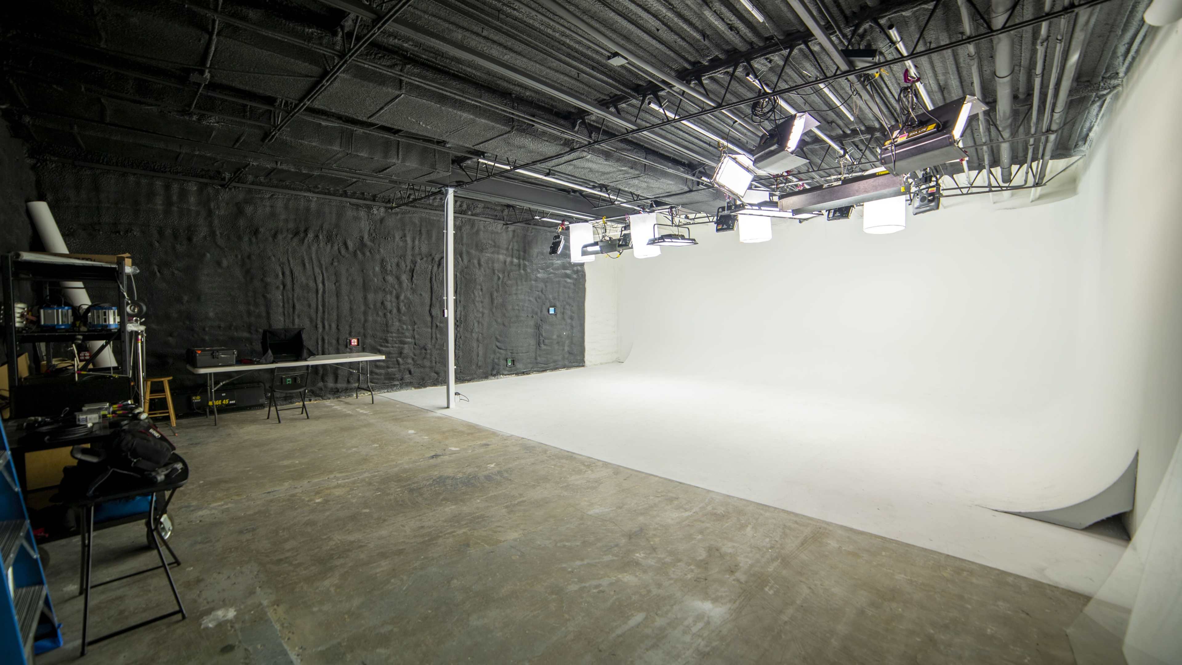 A spacious, empty photography studio with a white backdrop, overhead lights, and a table set against a dark wall.