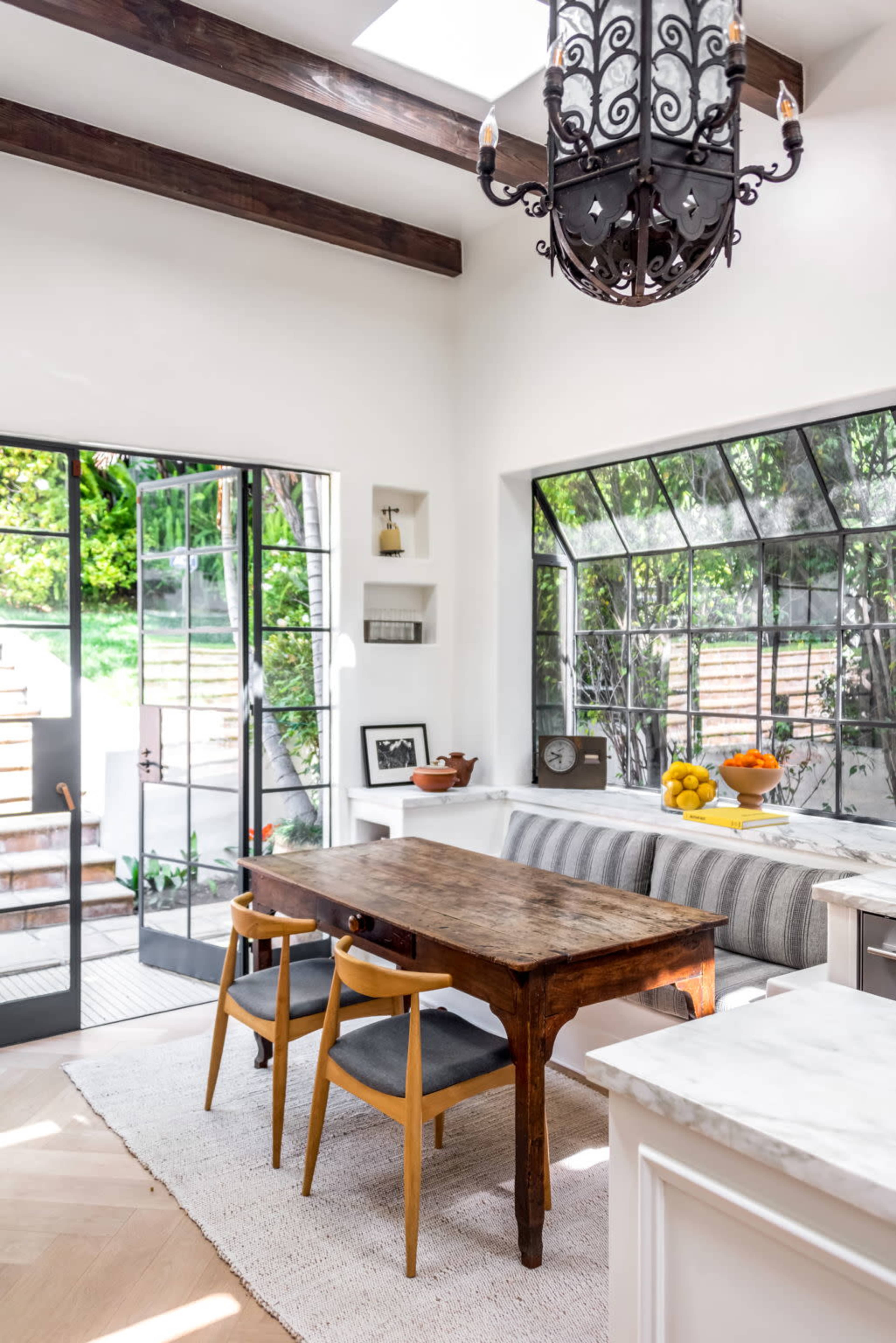 A dining area features a wooden table with chairs, a large window overlooking a garden, and a chandelier hanging from the ceiling.