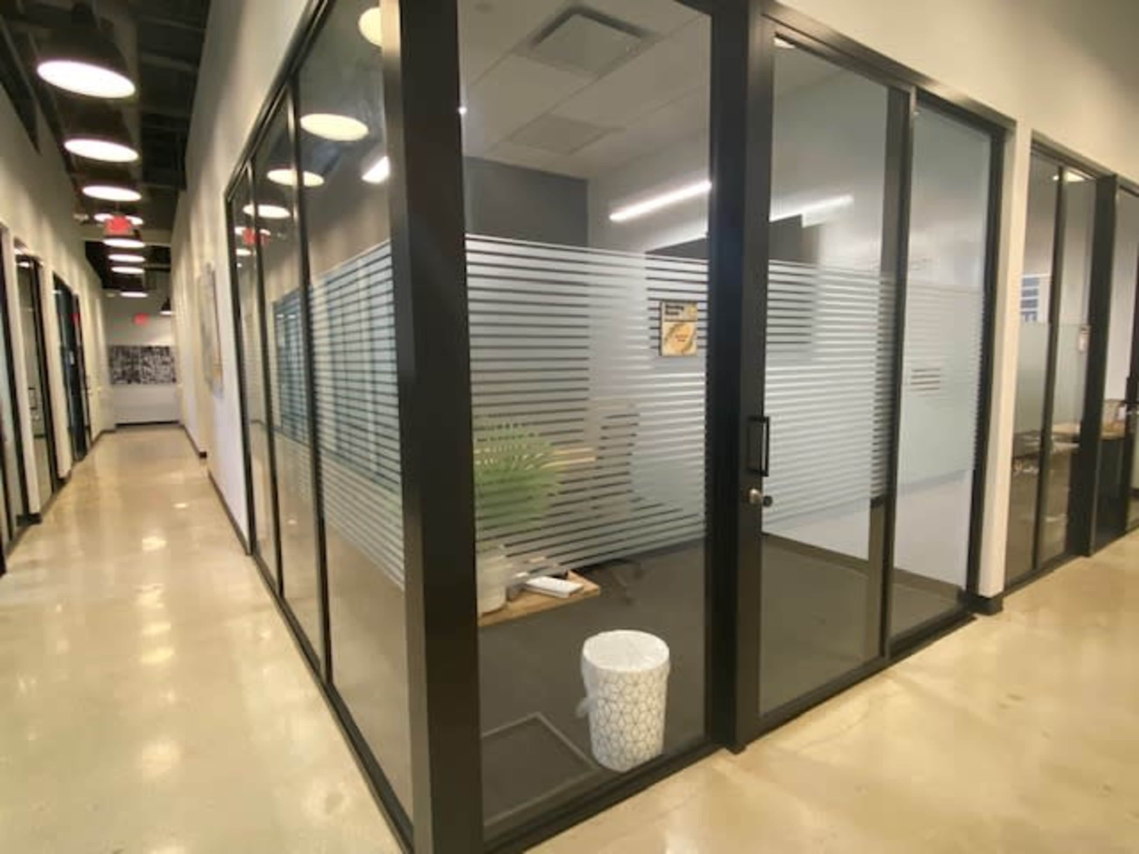 The image shows a modern glass-walled office with a small plant and a wastebasket, located in a corridor with overhead lighting.