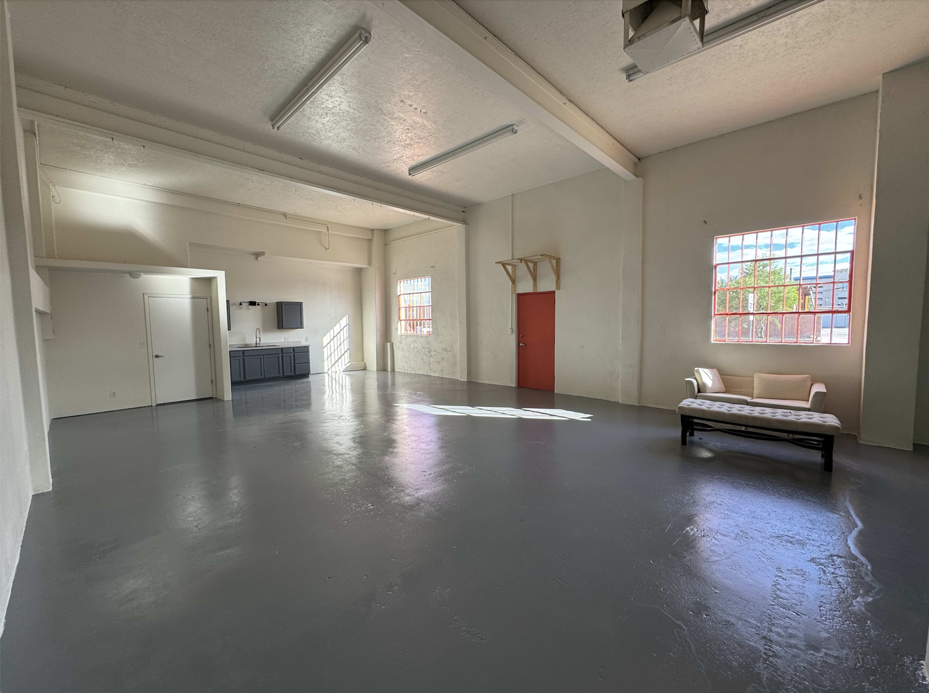The image shows a spacious, empty room with grey flooring, high ceilings, a small kitchen area in the back, and a single couch positioned in the foreground.