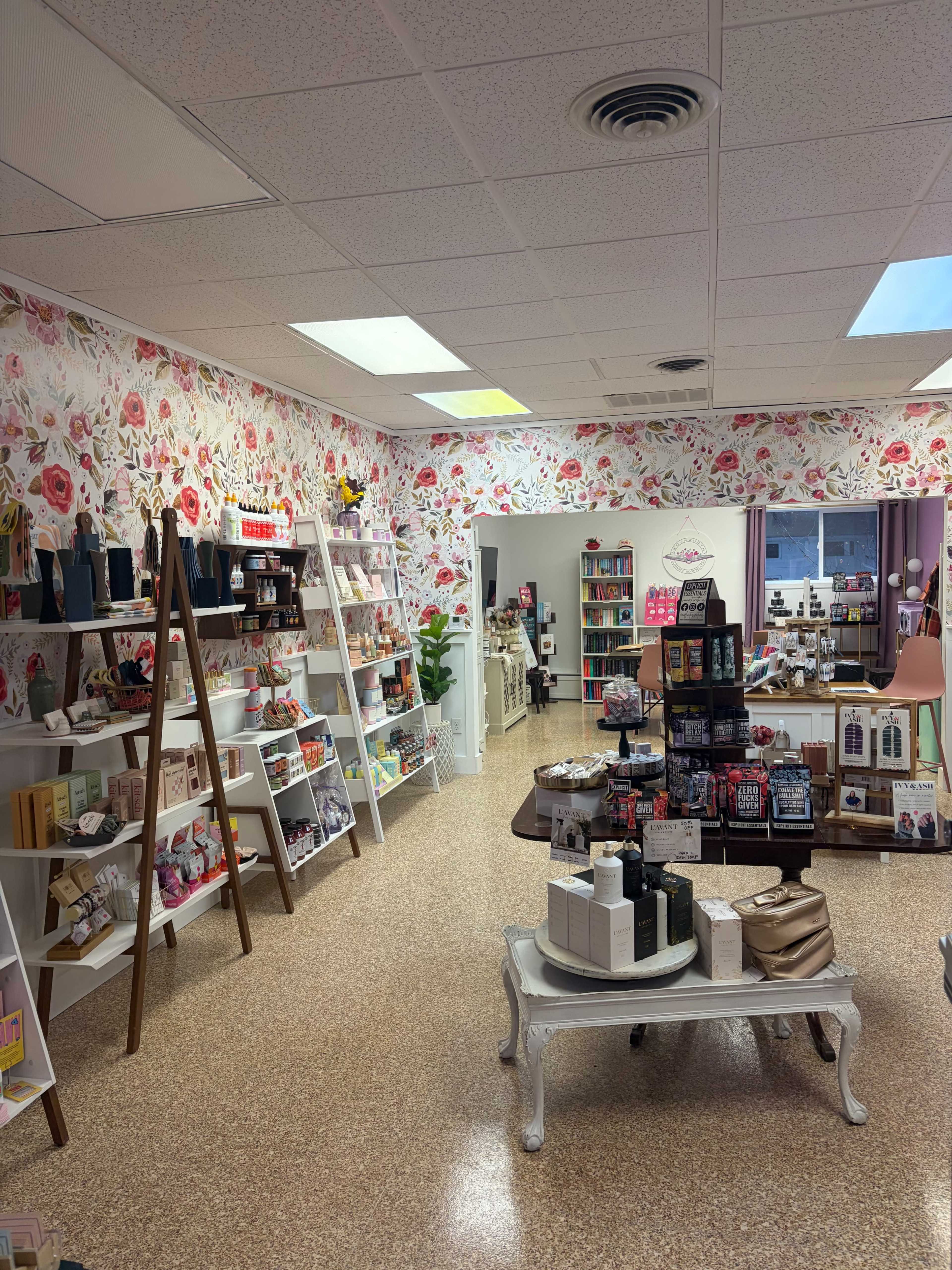 The image shows a brightly lit retail shop with floral wallpaper featuring shelves filled with various beauty and lifestyle products, and a central display table with additional items for sale.