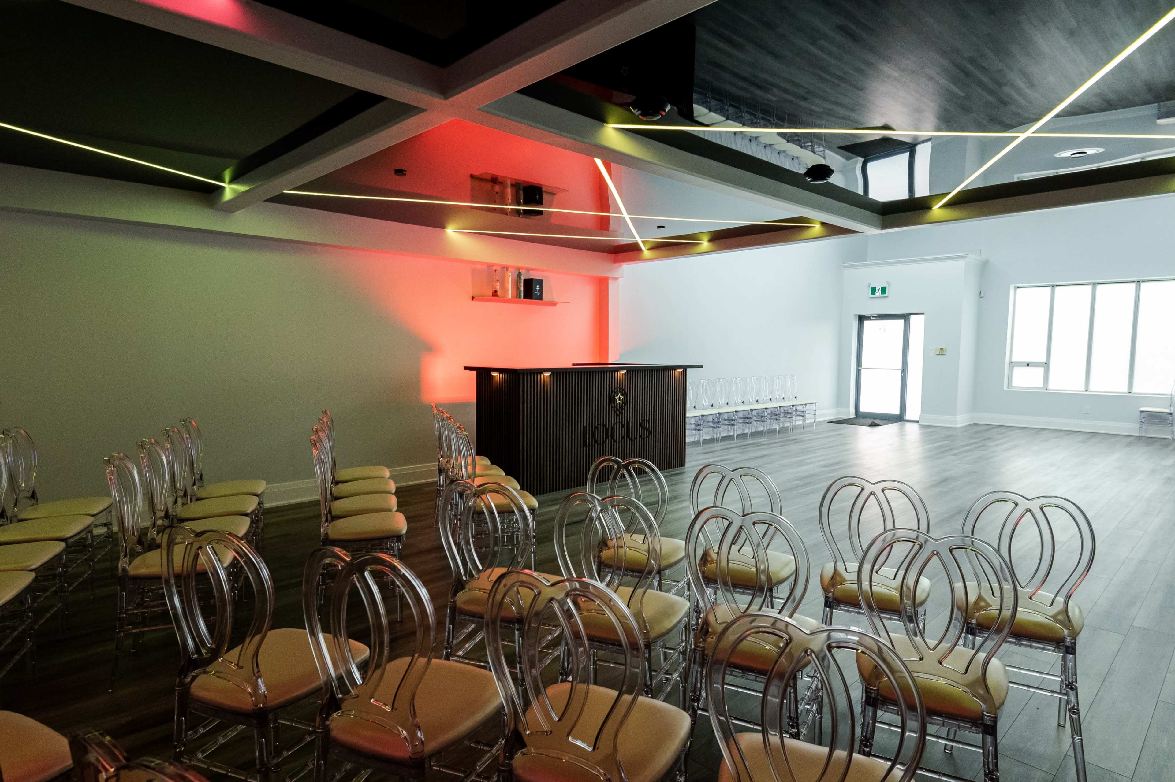 A conference room features rows of acrylic chairs facing a wooden bar with backlighting, set against a modern ceiling design.