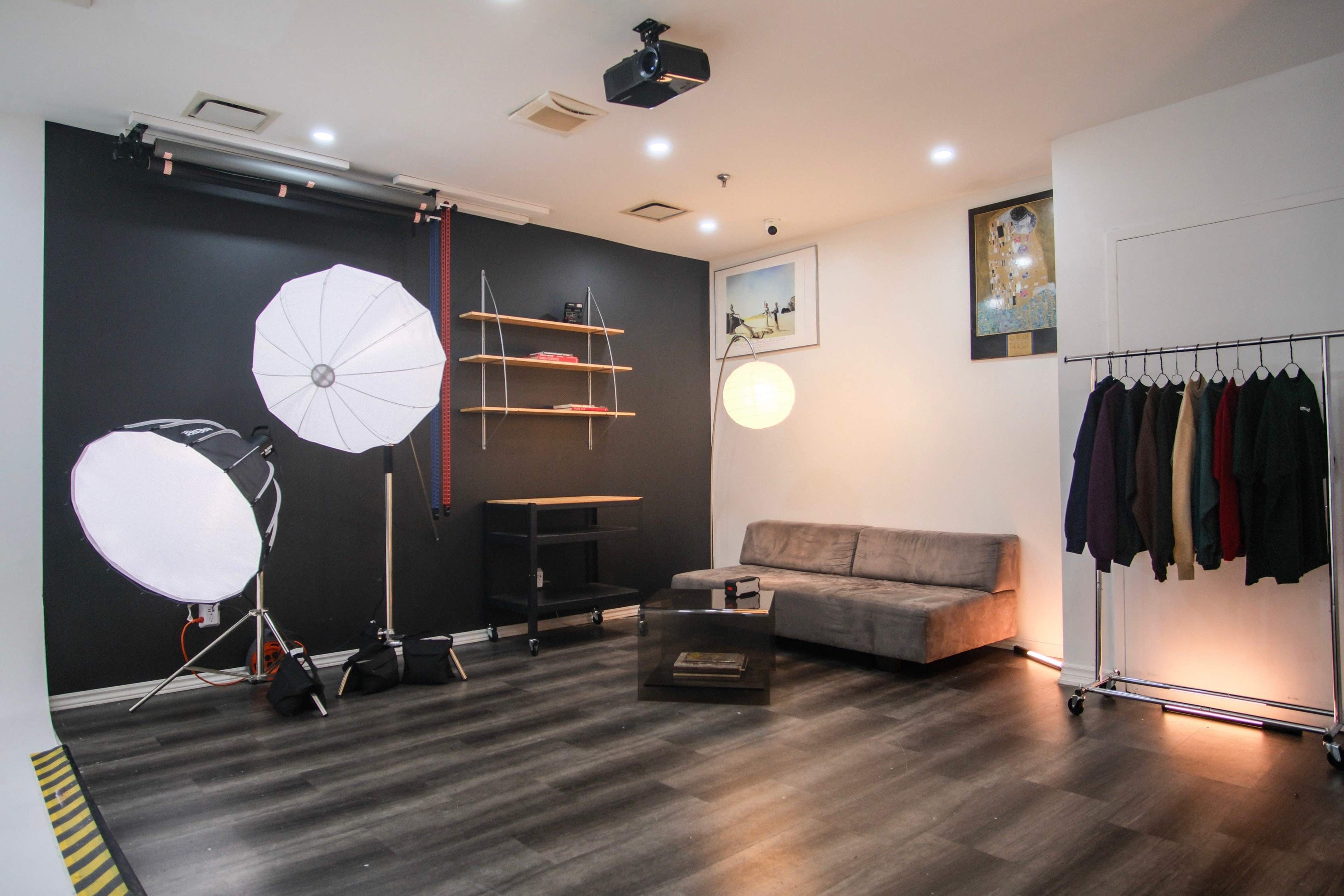 A studio setting with lighting equipment, a gray sofa, a table, shelves, and a clothing rack against a dark wall.