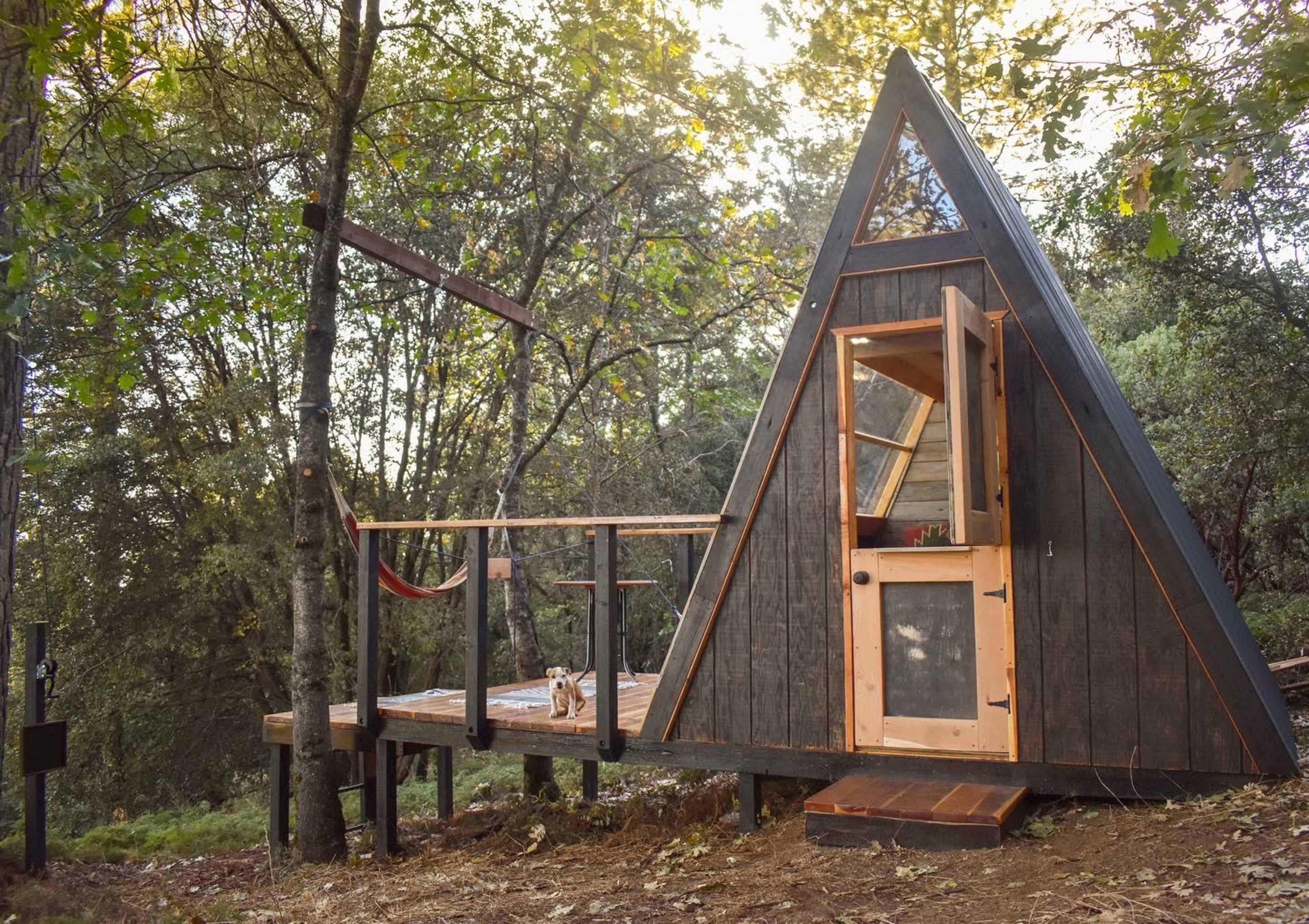 A triangular wooden cabin with an overhanging deck is nestled among trees in a forested setting.
