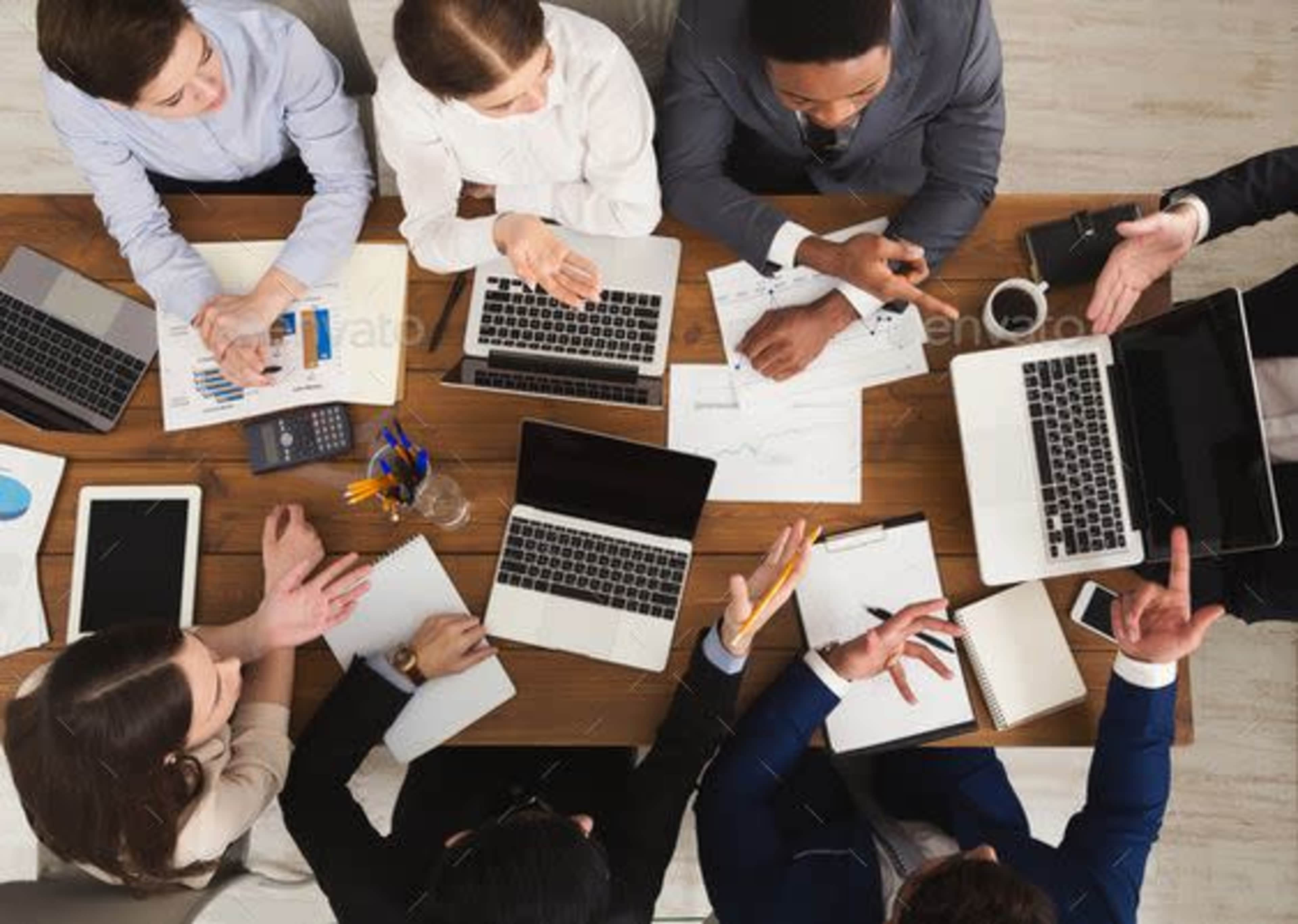 A group of professionals is engaged in a meeting around a wooden table with laptops, documents, and office supplies.