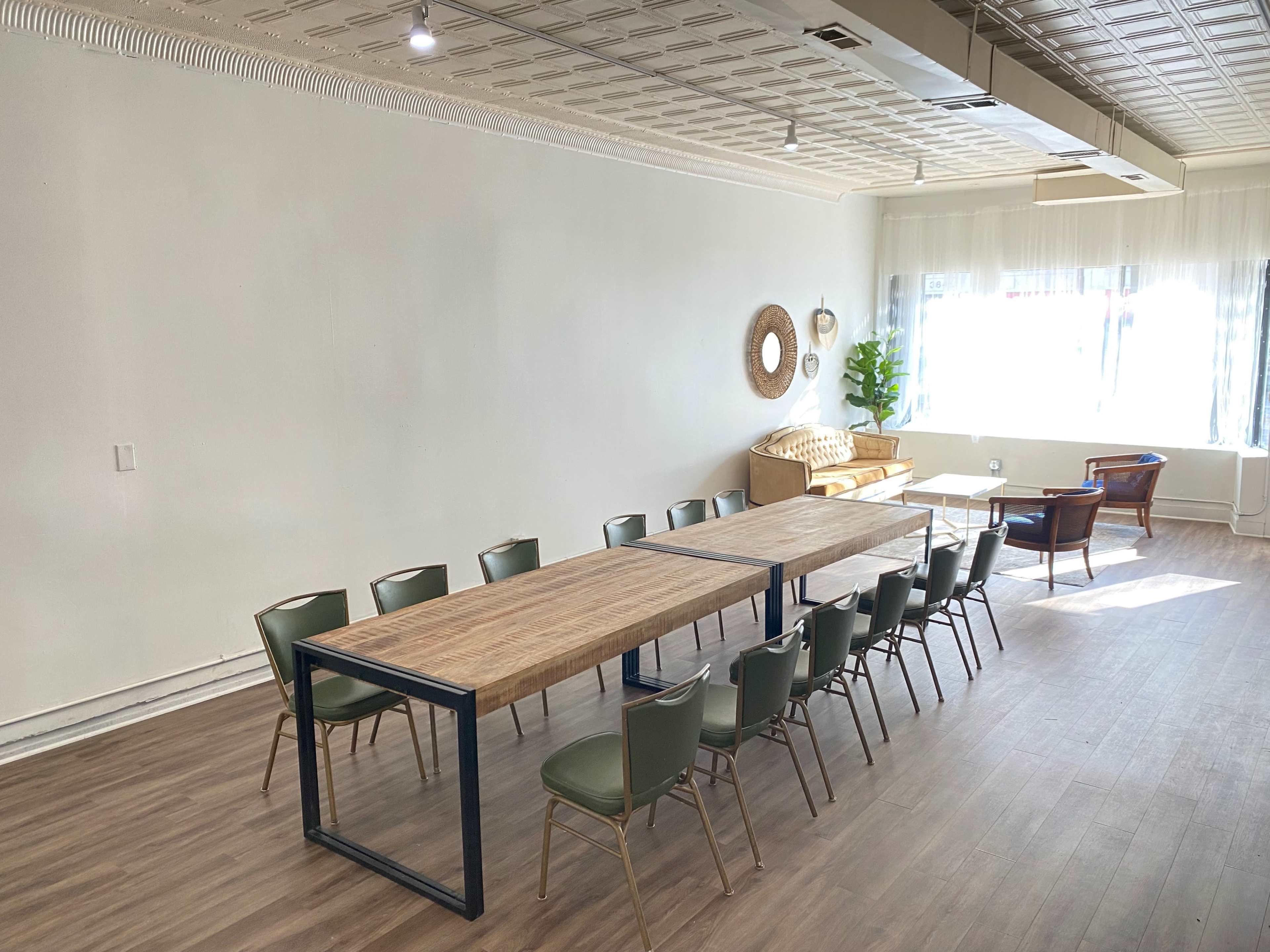 A spacious room with a long wooden table surrounded by chairs, a sofa in the corner, and large windows letting in natural light.