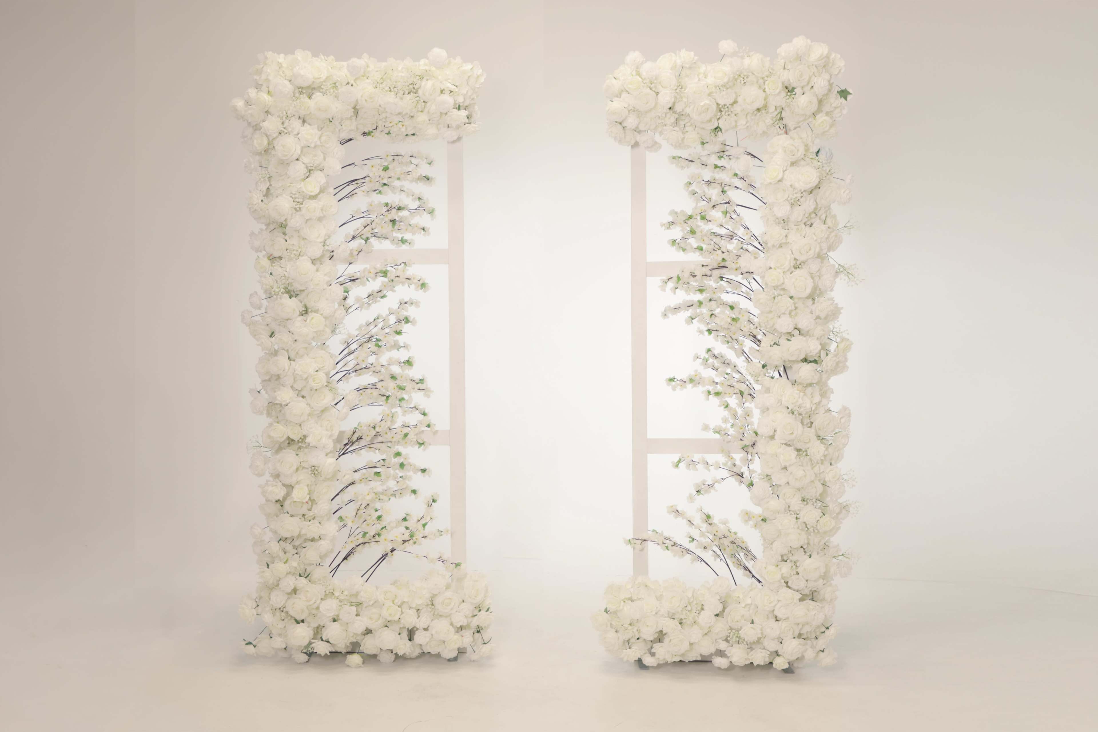 Two tall, white floral archways are positioned parallel to each other, adorned with a variety of white flowers and greenery.