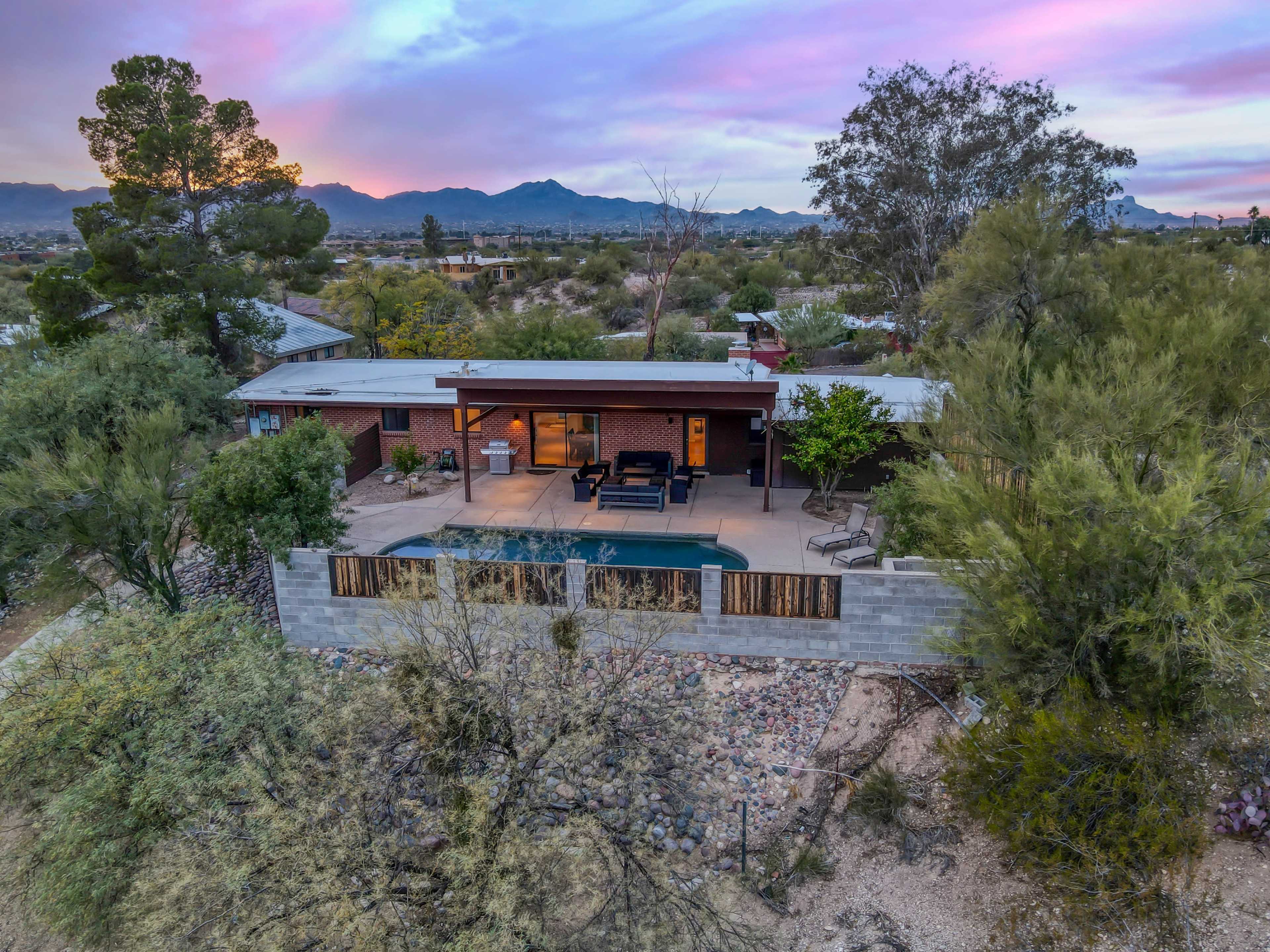 A single-story brick house with a patio and pool is surrounded by desert vegetation and mountains in the background.