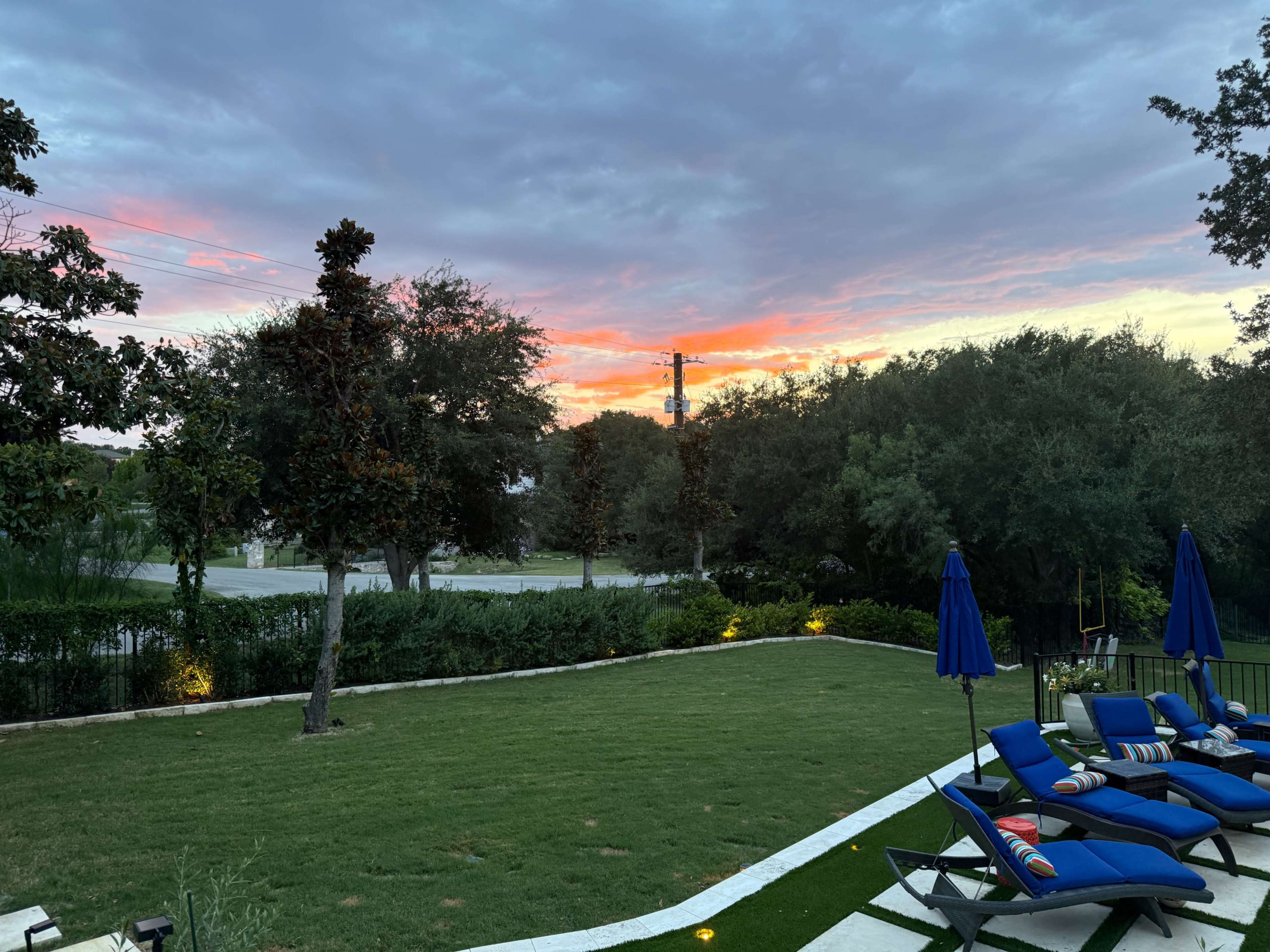 The image depicts a backyard with lounge chairs overlooking a sunset with pink and orange hues, framed by trees and a landscaped lawn.