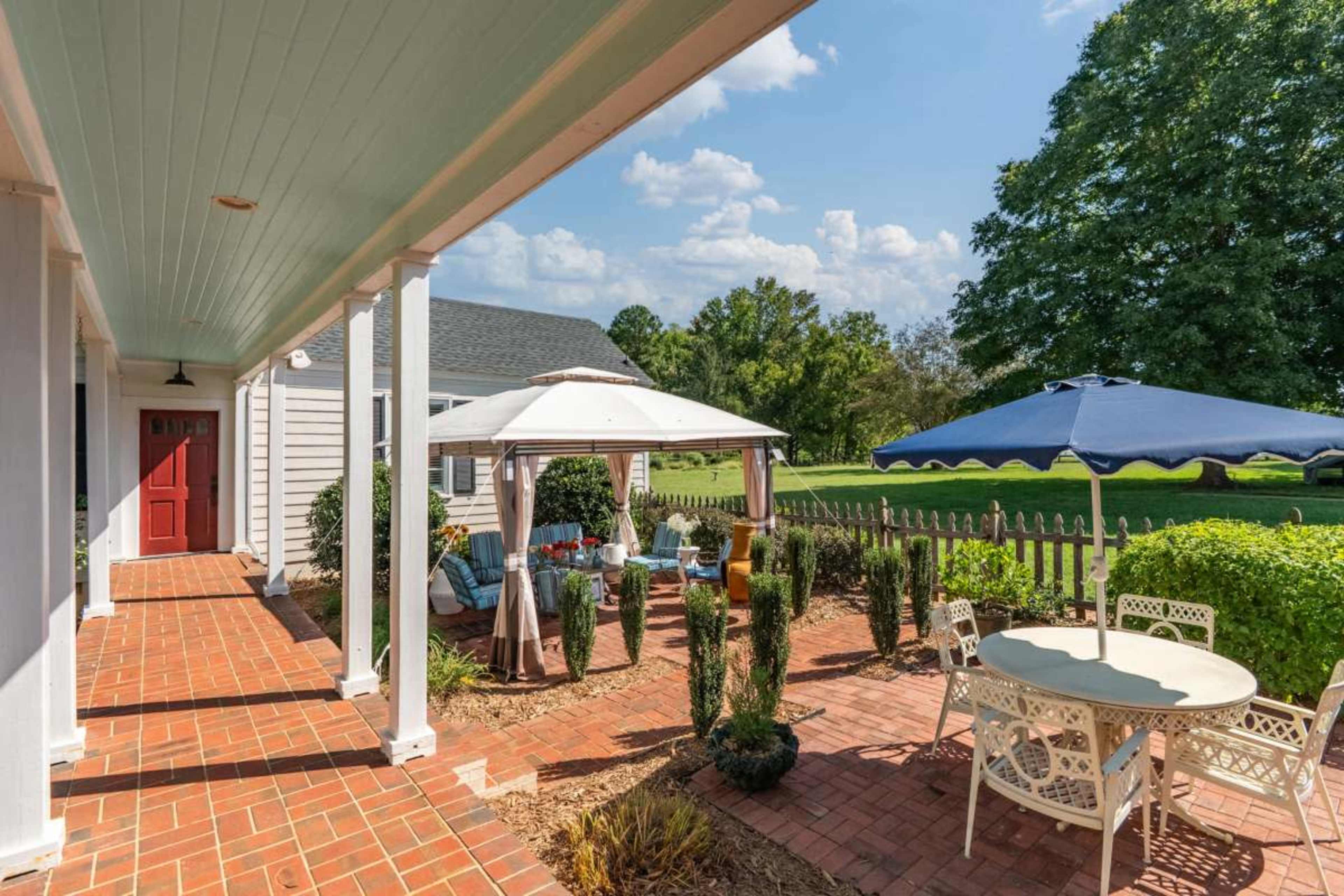 The scene shows a brick patio with outdoor furniture, shaded by large umbrellas, surrounded by greenery and a wooden fence.