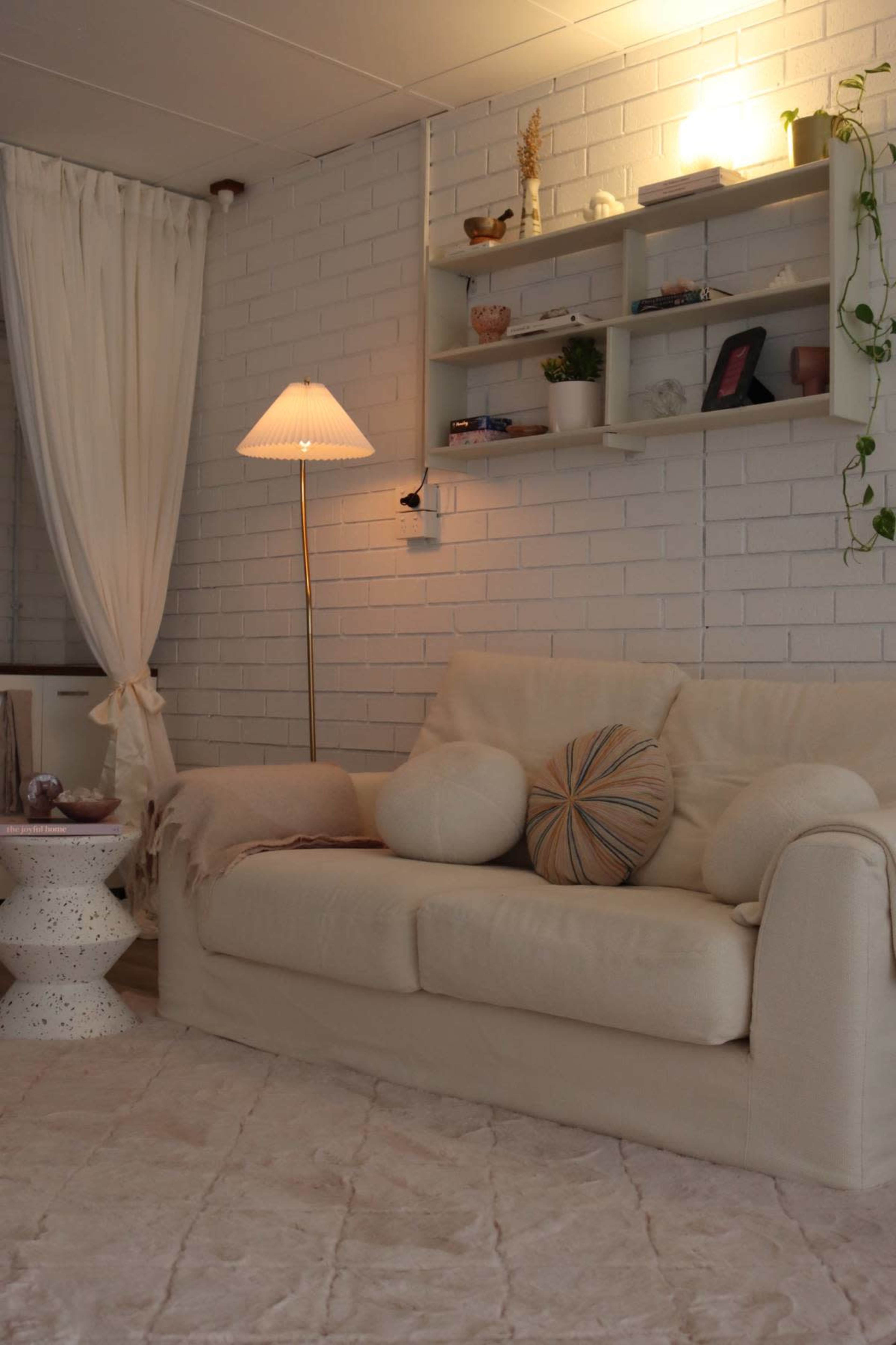 The image shows a cozy living room featuring a white sofa with decorative pillows, a small round side table, and a lamp, all set against a brick wall and a shelf with various decor items.