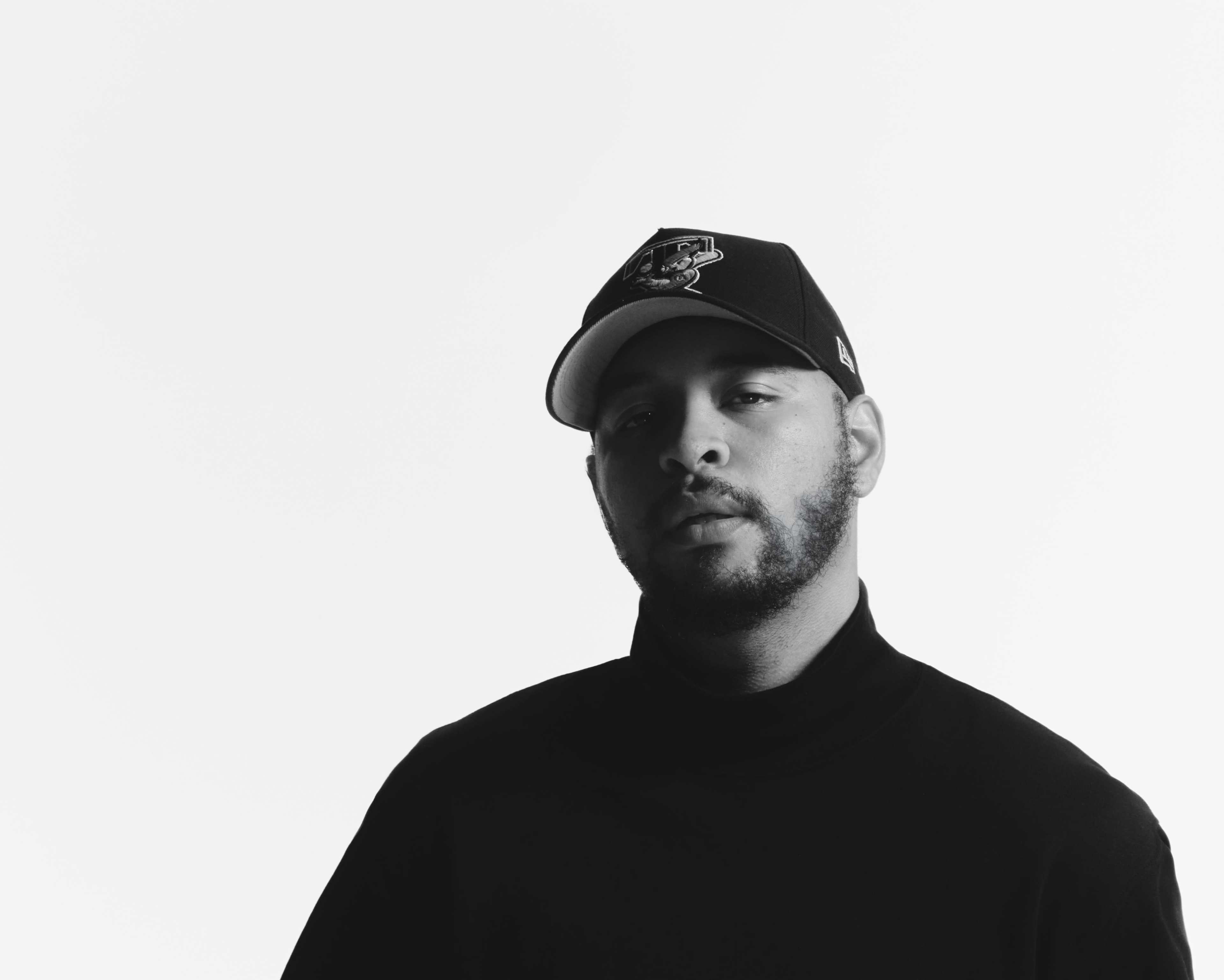 A man wearing a black turtleneck and a baseball cap poses against a white background.