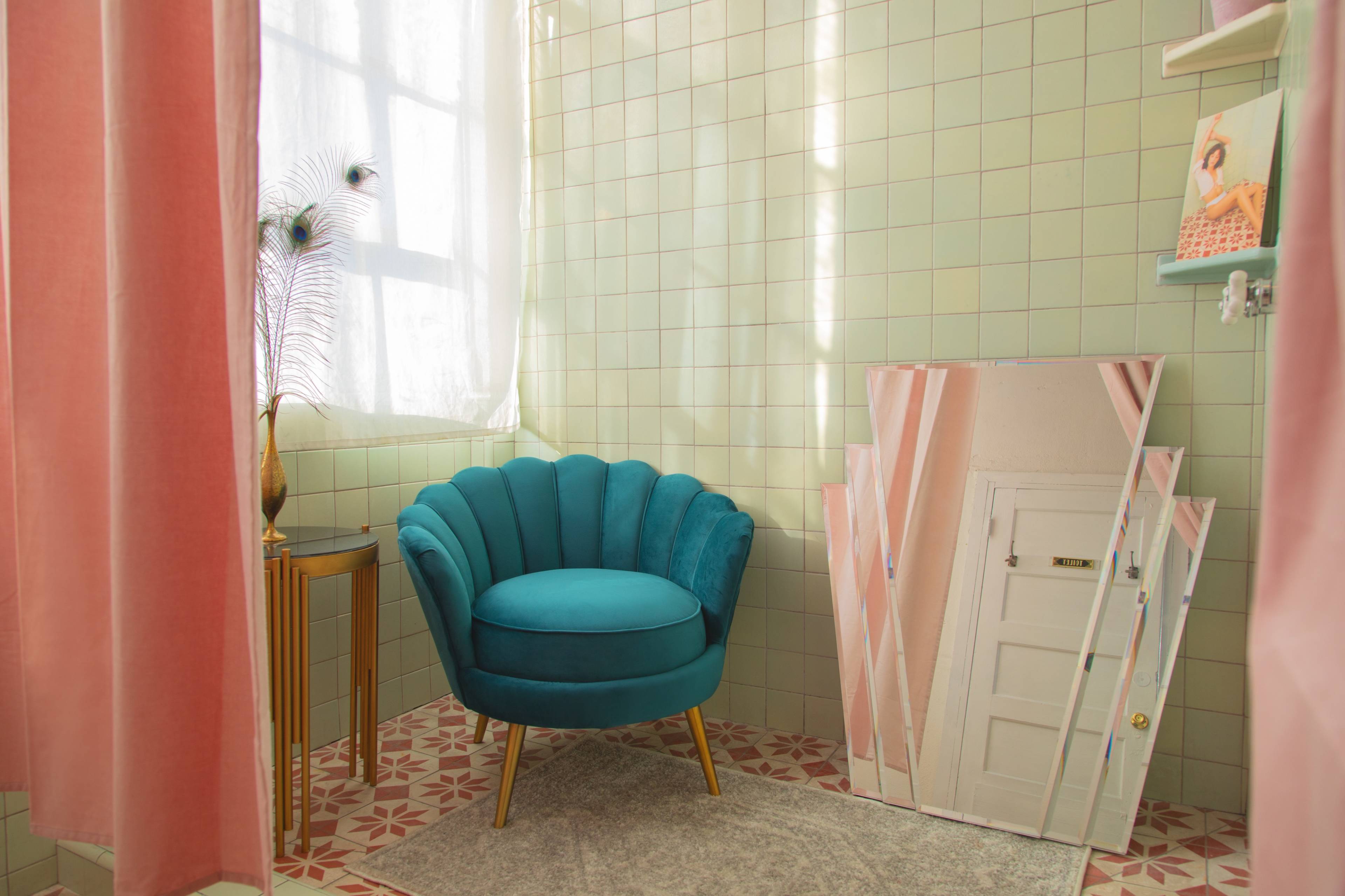A styled corner of a bathroom featuring a teal armchair, a small gold side table, and a distorted mirror against a green tiled wall.