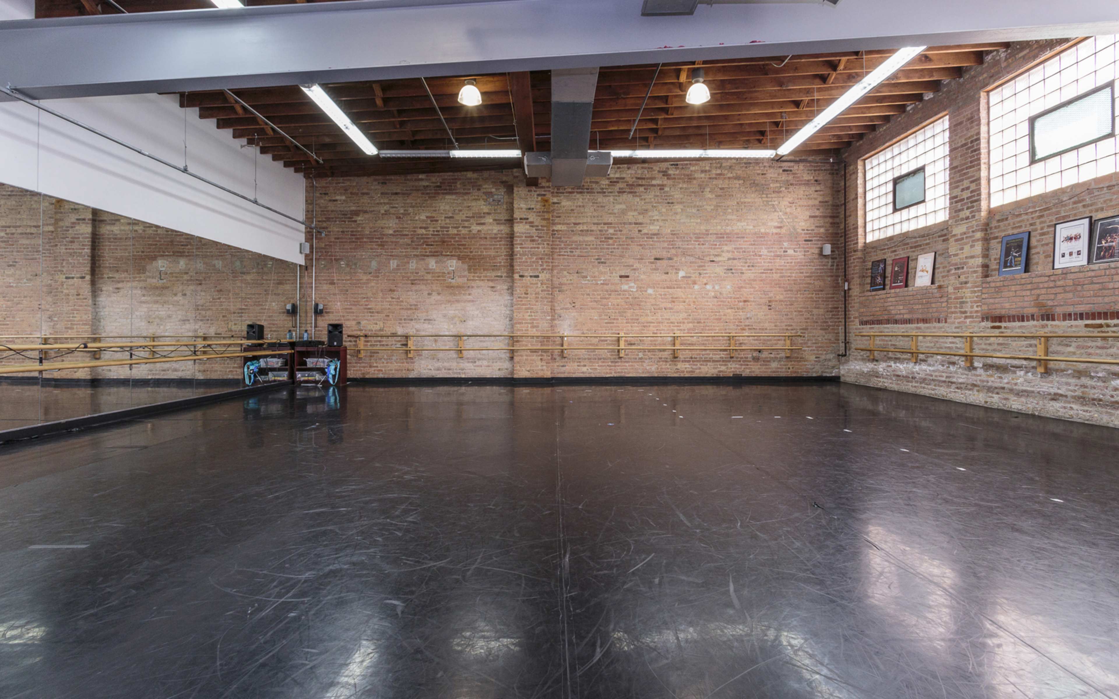 A spacious dance studio features a polished black floor, exposed brick walls, and large windows allowing natural light.