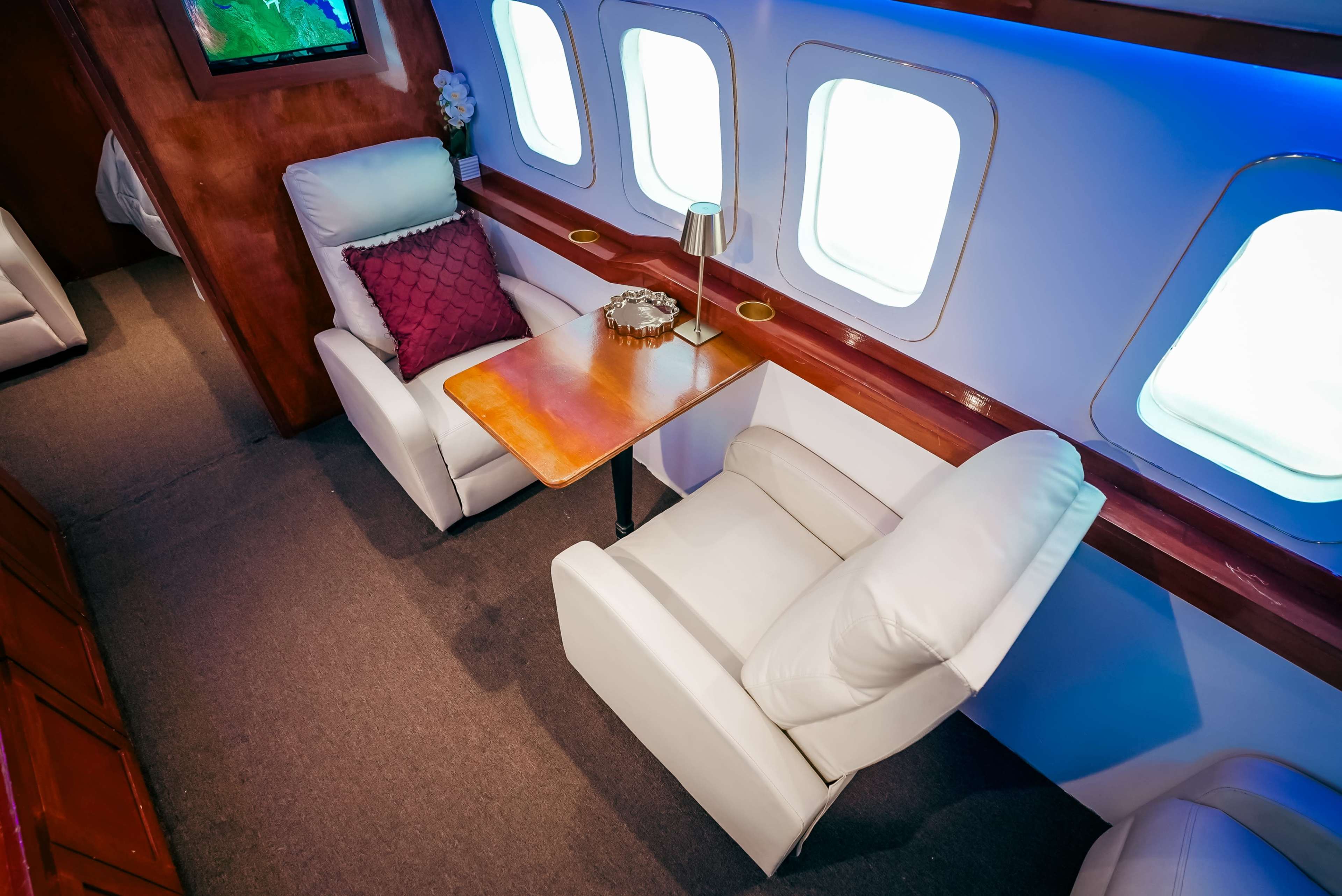The image shows two white armchairs beside a wooden table in a modern aircraft interior with large windows.