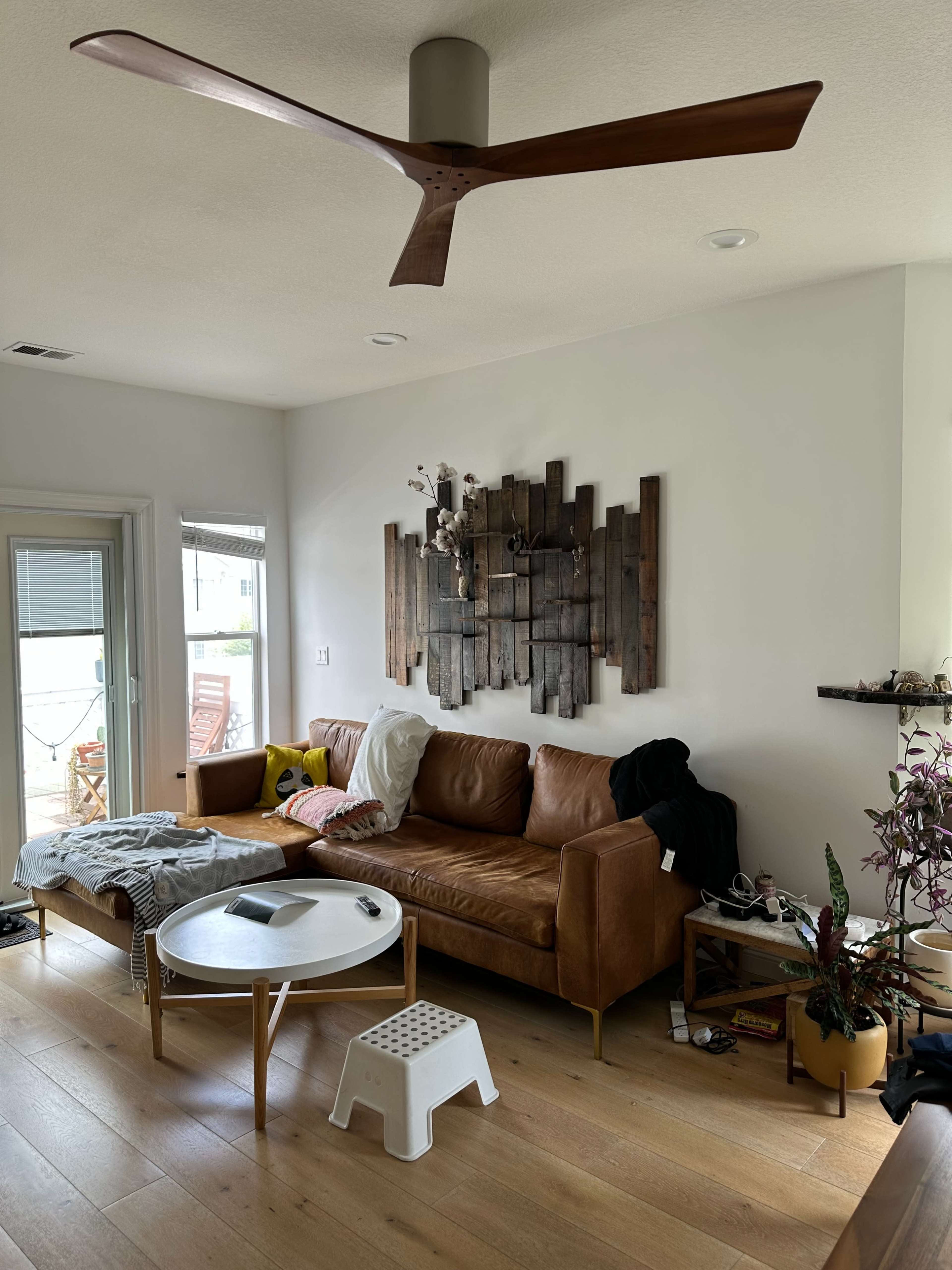 The living room features a brown leather couch, a round coffee table, a wooden wall art installation, and a ceiling fan.