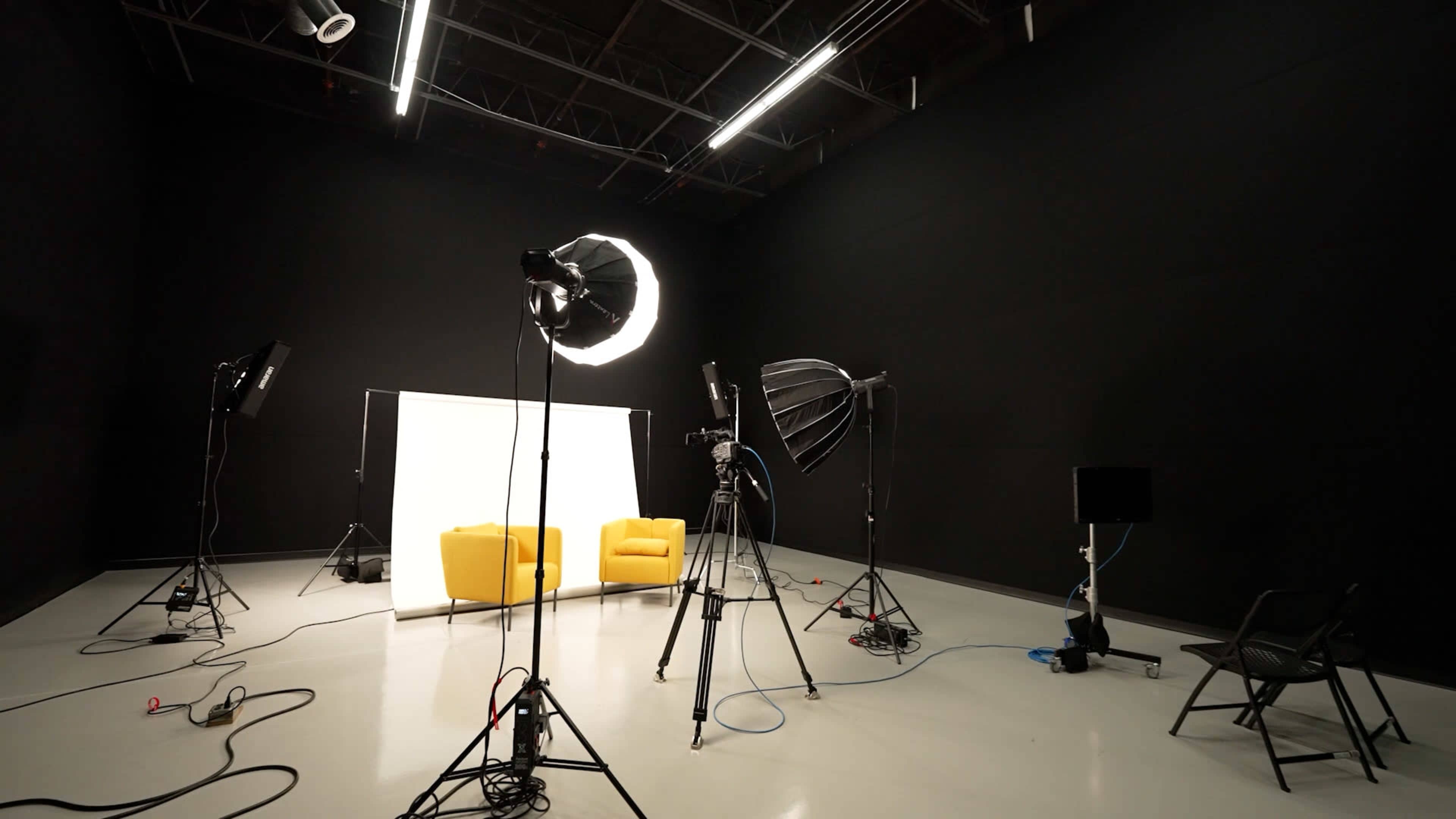 A dark studio space set up for a photoshoot, featuring two yellow chairs, lighting equipment, and a backdrop.