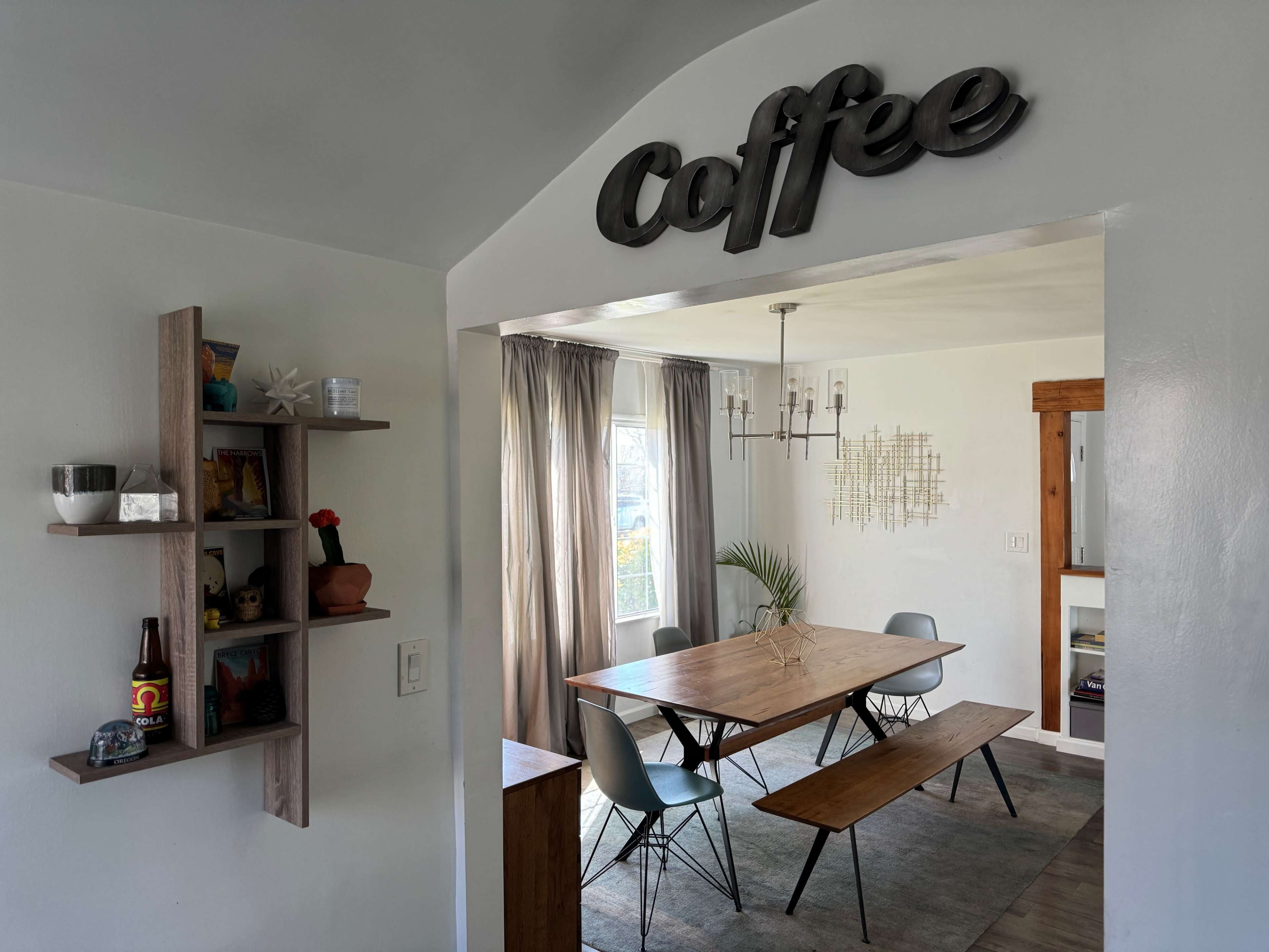 A dining area features a wooden table surrounded by chairs, a coffee-themed wall sign, and decorative shelves.
