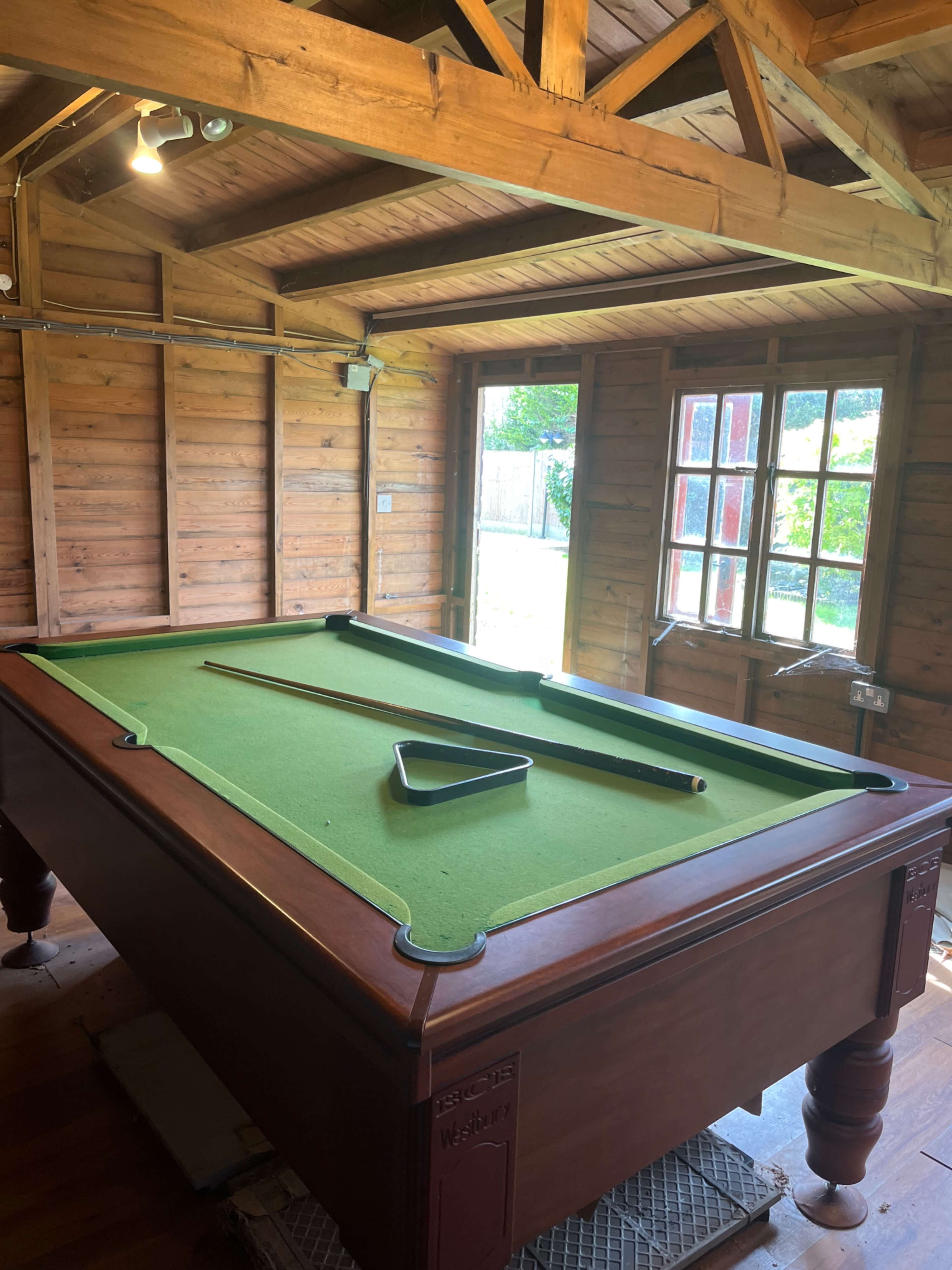 The image shows a wooden pool table with a green felt surface inside a rustic wooden room with a window overlooking a backyard.