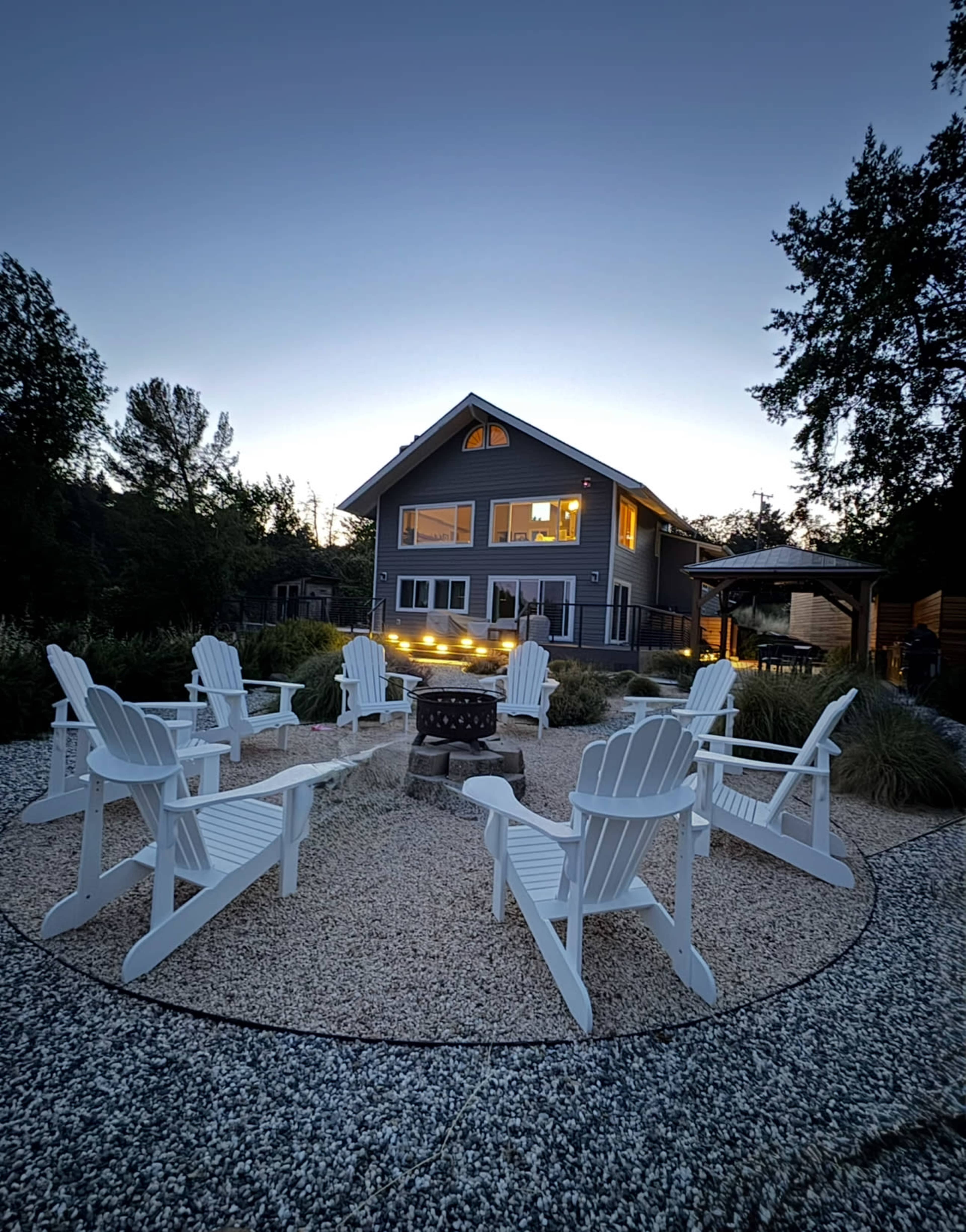 A circular arrangement of white Adirondack chairs surrounds a fire pit in a gravel area, with a two-story house and trees in the background as dusk approaches.