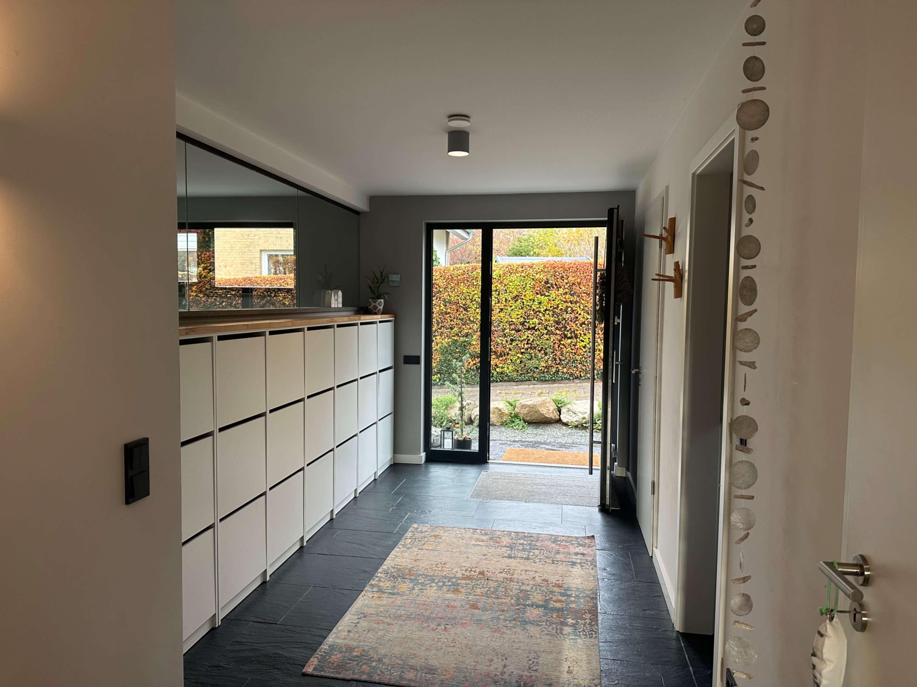 The image shows a modern hallway featuring a white storage unit, a patterned rug, and glass doors that provide a view of a garden.