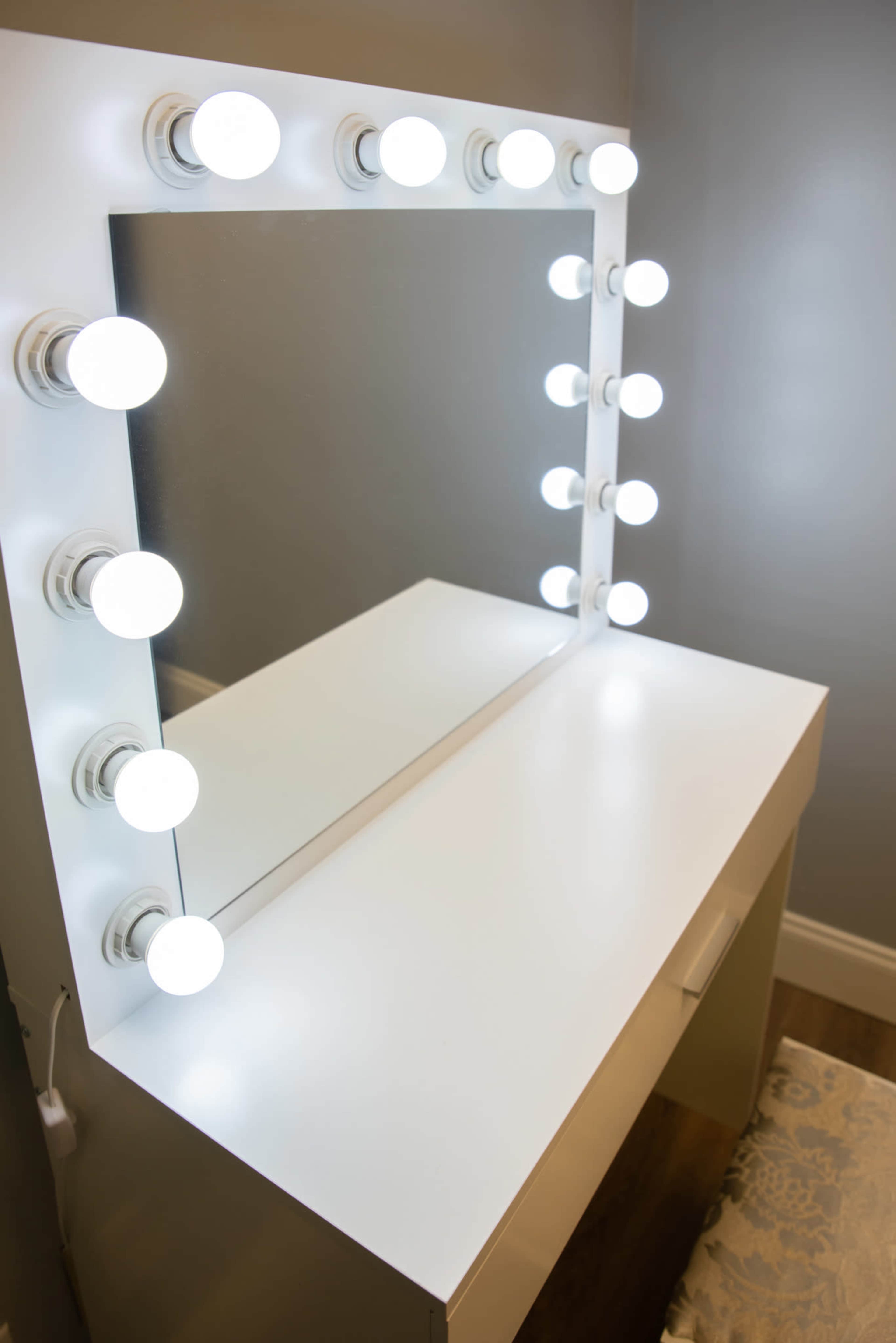 A white vanity with a large mirror surrounded by bright round light bulbs is positioned against a gray wall.