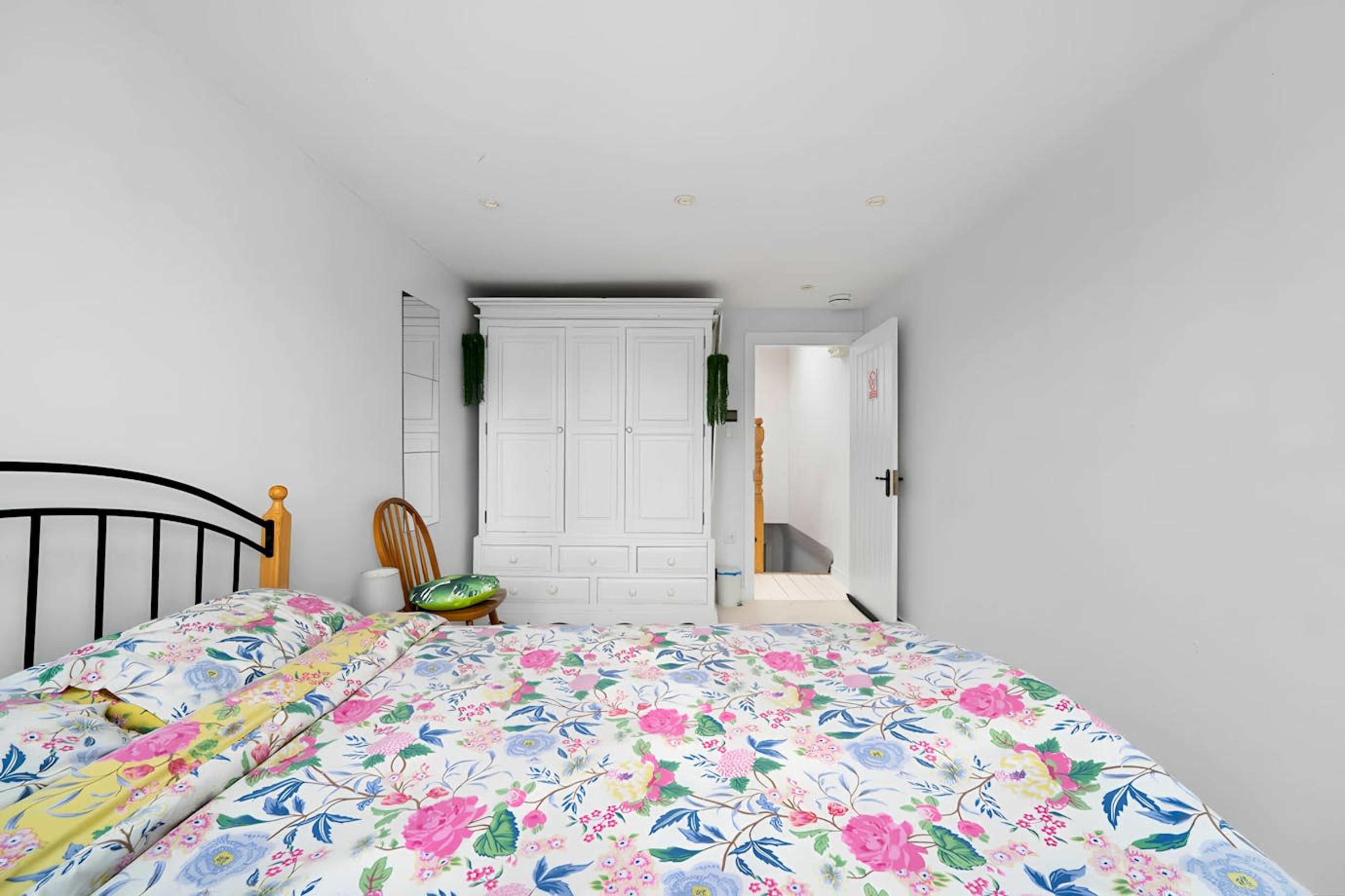 A cozy bedroom features a bed with a floral-patterned blanket and a white wardrobe, with a wooden chair and a glimpse of a hallway in the background.