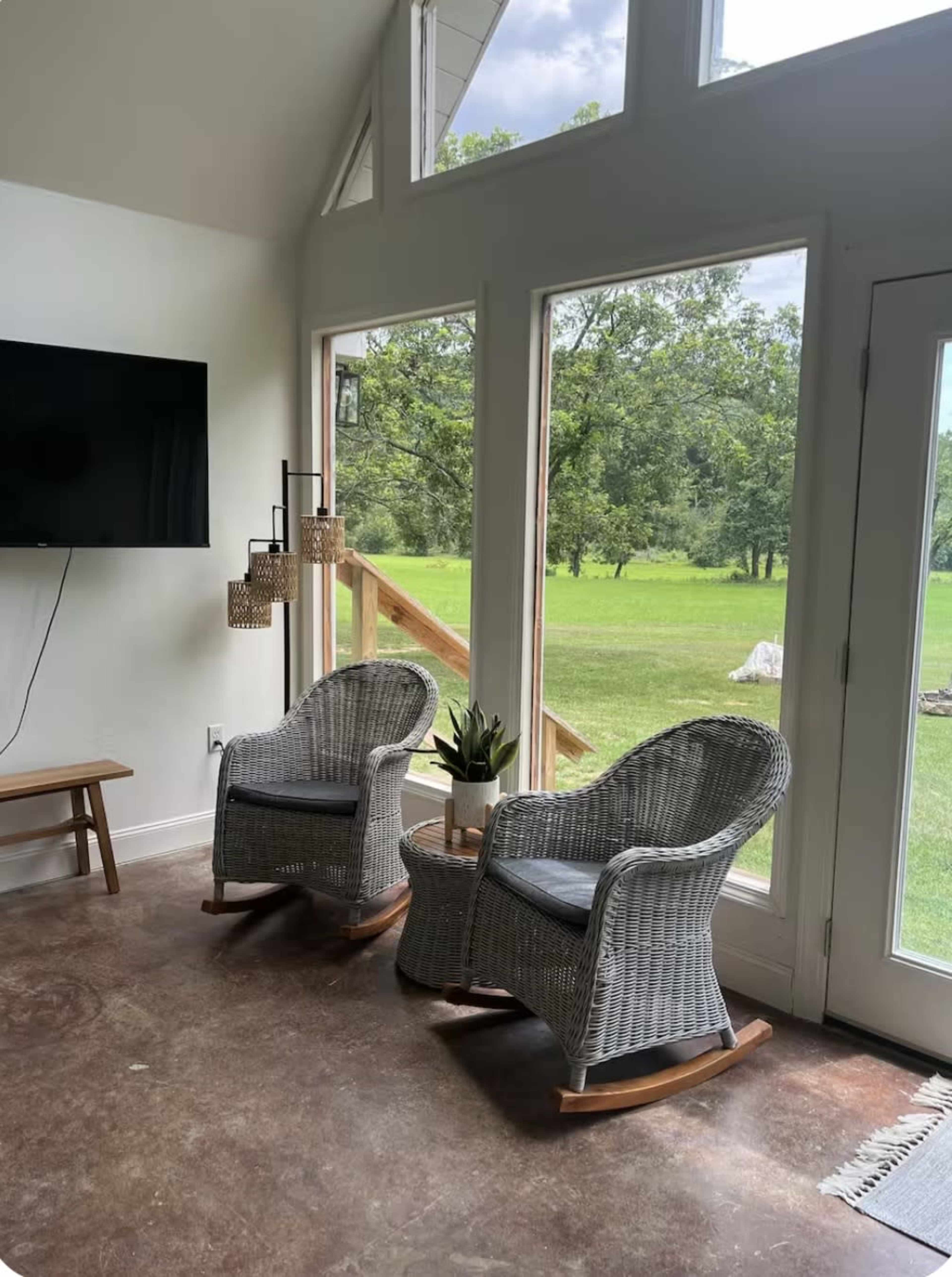 Two wicker rocking chairs are positioned by large windows that overlook a grassy outdoor area, with a small table and a plant nearby.