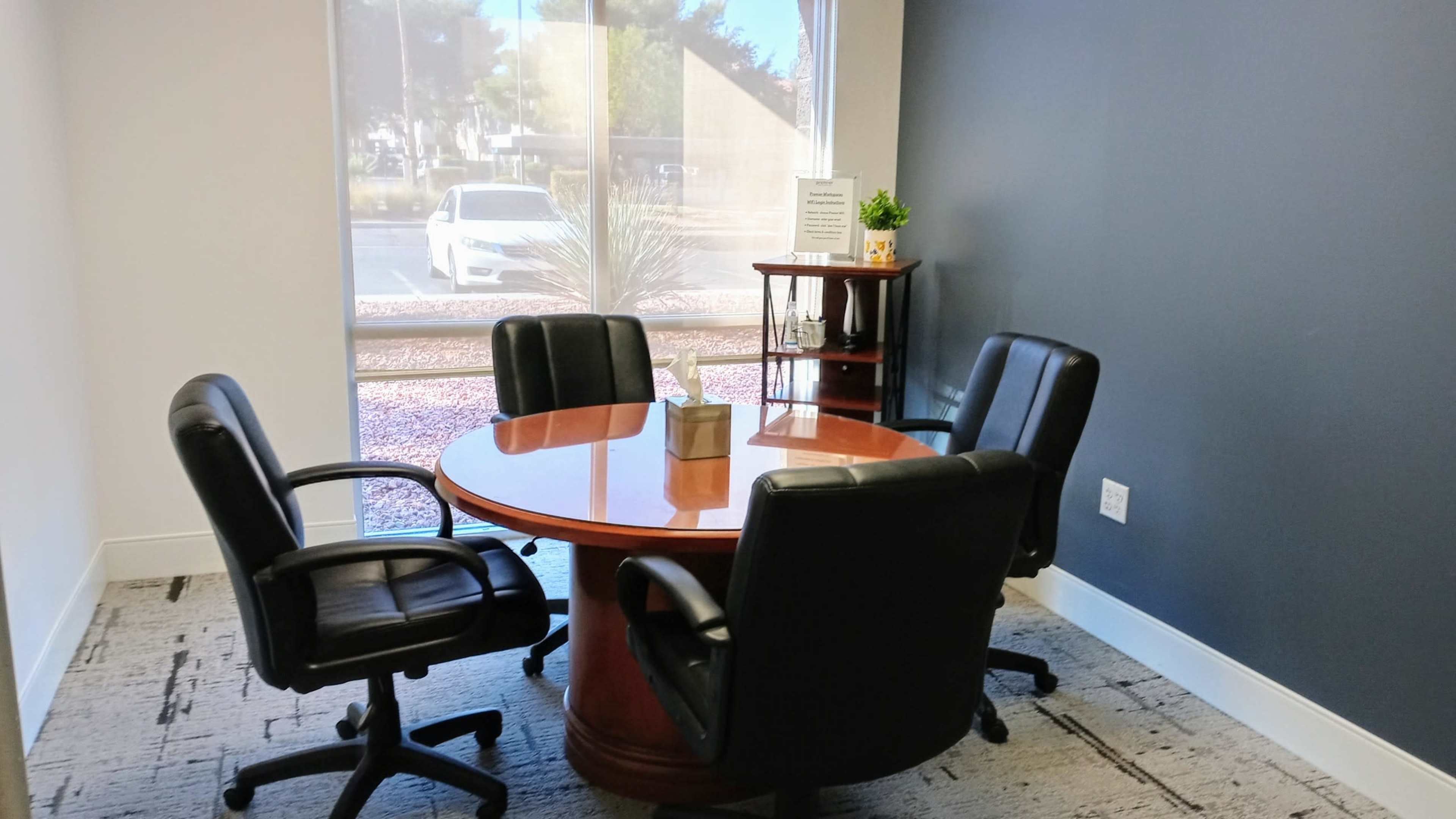 A round wooden table surrounded by four black office chairs is positioned in a small meeting room with a window facing an outdoor view.