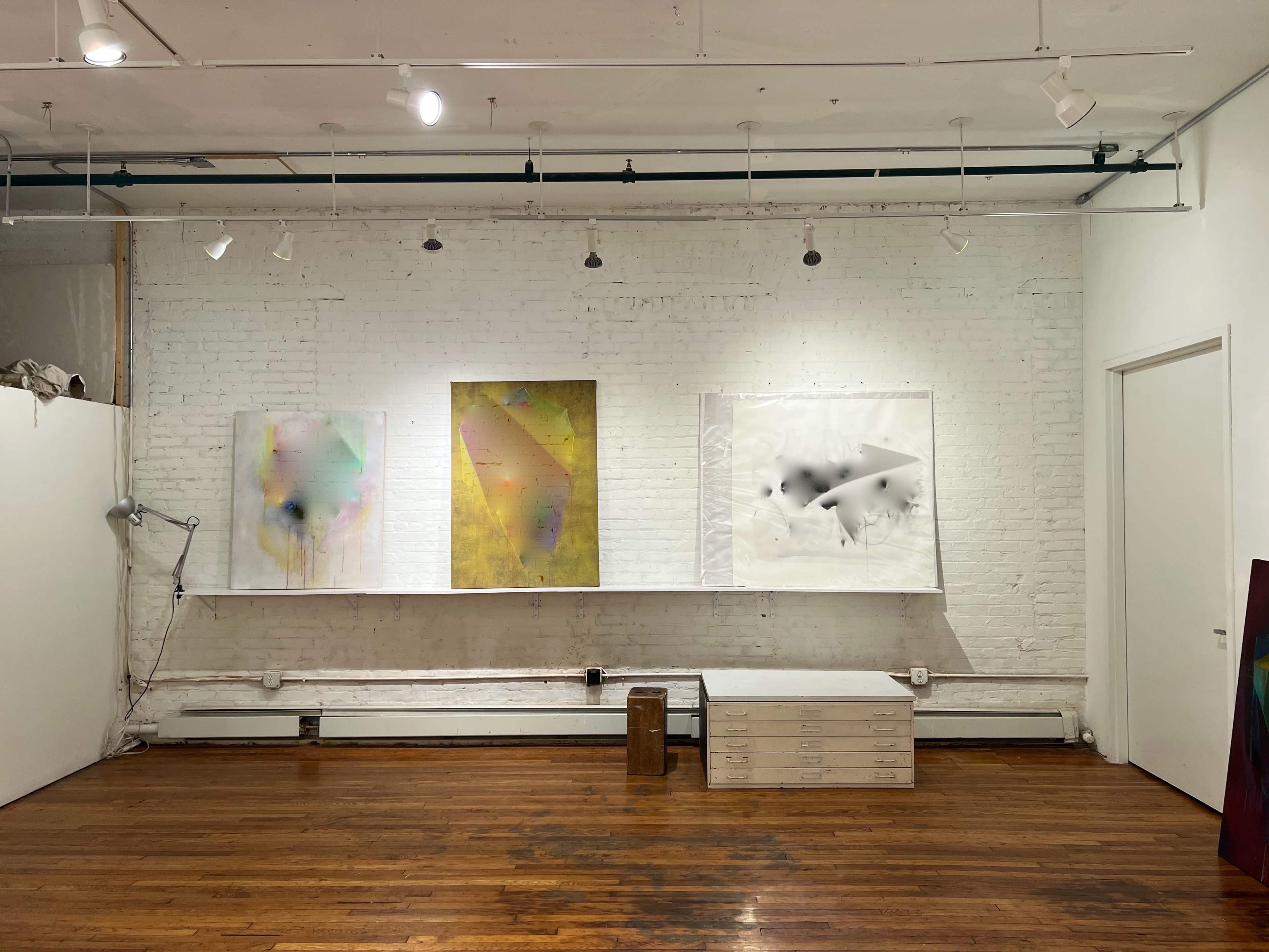 An art gallery with three abstract paintings displayed against a white brick wall, complemented by wooden flooring and overhead lighting.