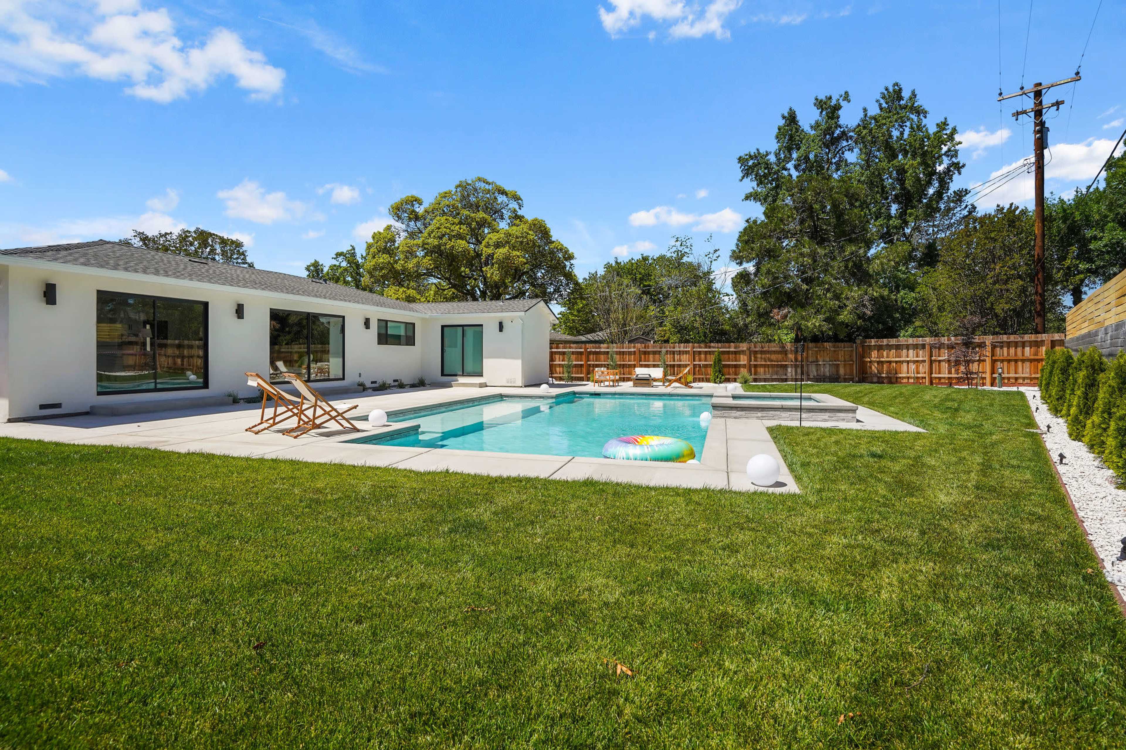 The image shows a modern backyard with a swimming pool, lounge chairs, and a well-maintained lawn surrounded by a wooden fence.