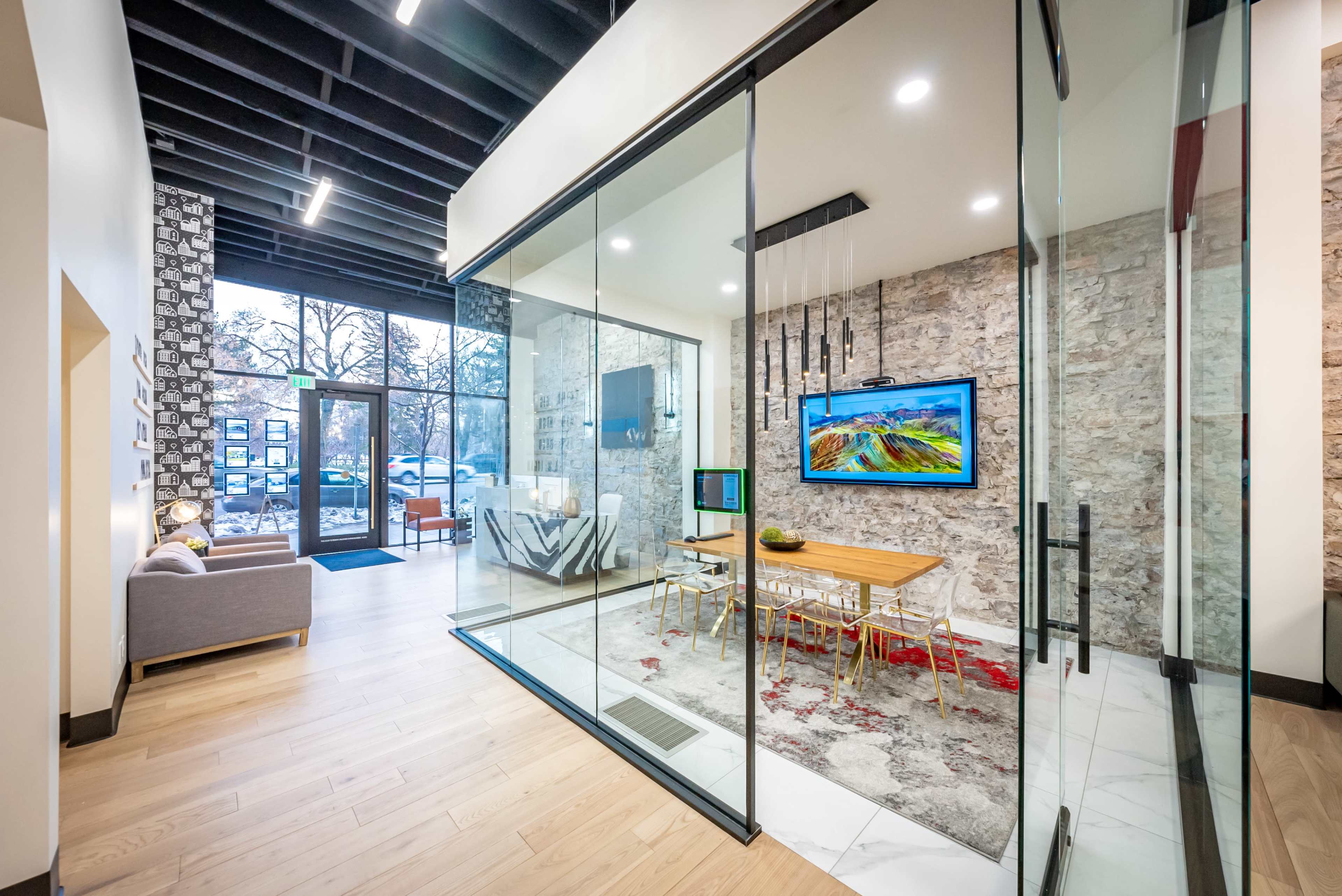 A modern office space features a glass-enclosed conference room with a dining table and a large TV, adjacent to an open reception area with a cozy seating arrangement.