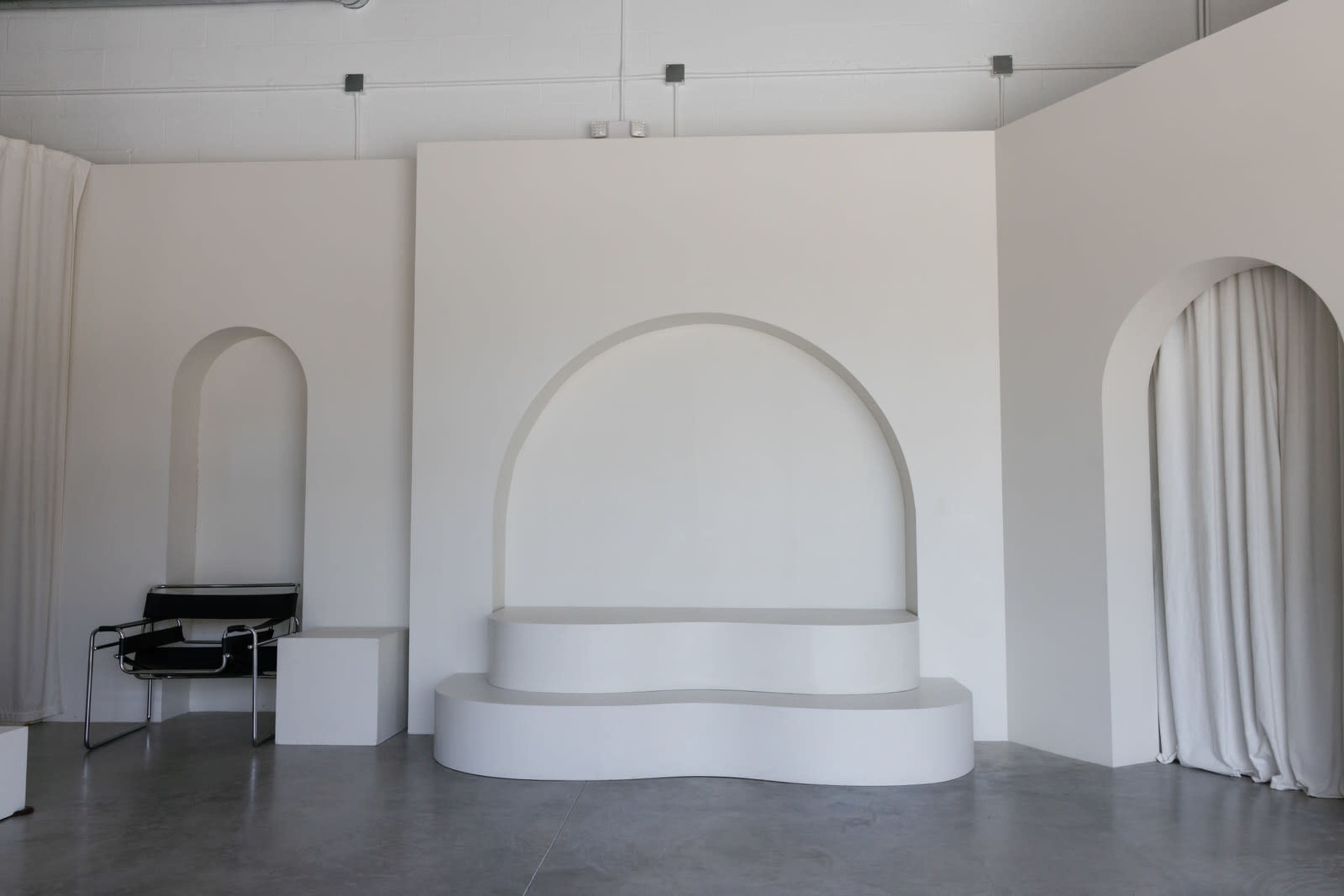 A minimalist interior space featuring curved architectural elements, a sectional seating area, and a black chair against a white backdrop.