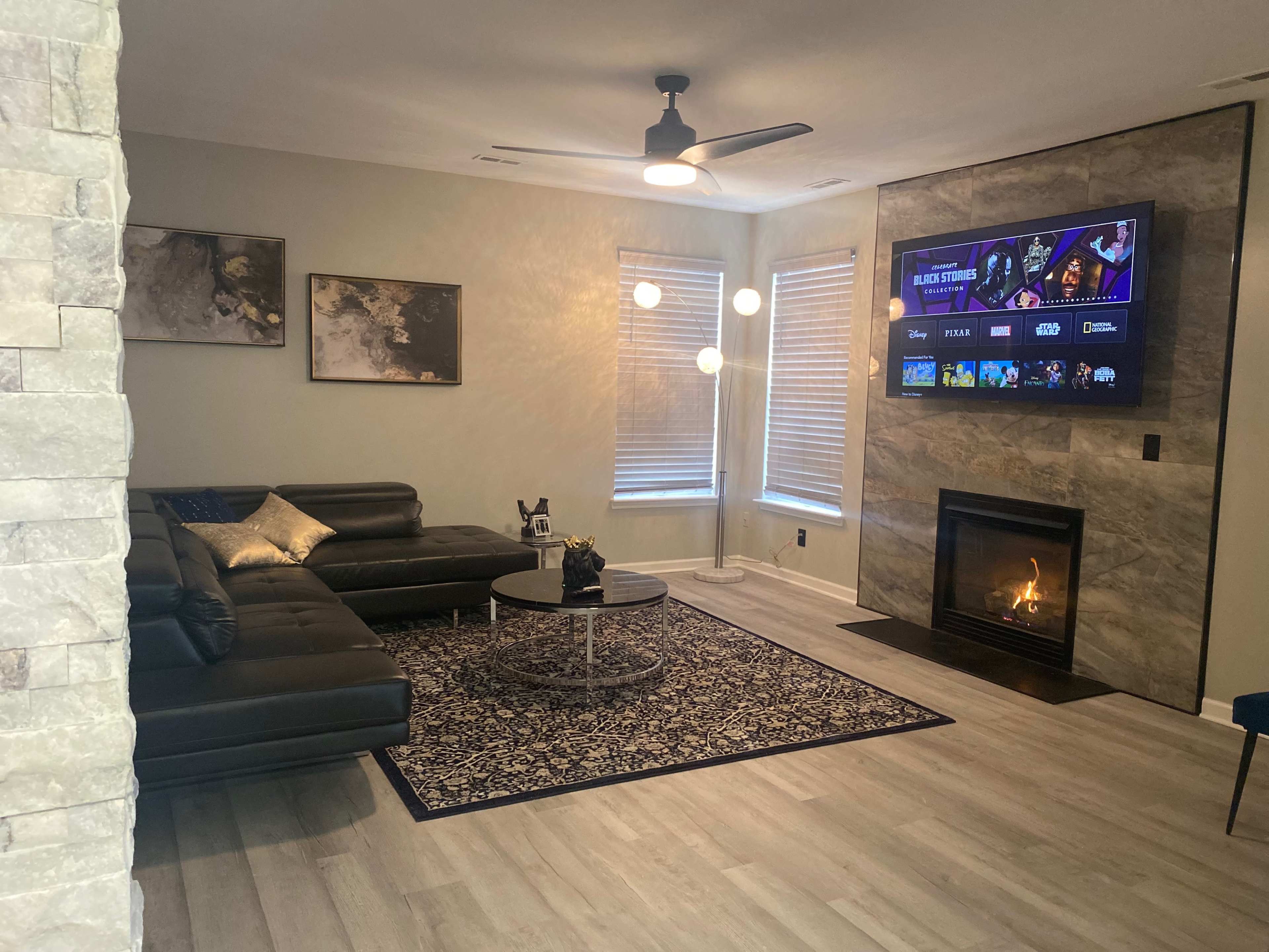 The image shows a modern living room with a sectional sofa, a circular coffee table, a large wall-mounted TV, and a fireplace beneath it, all arranged on light-colored flooring.