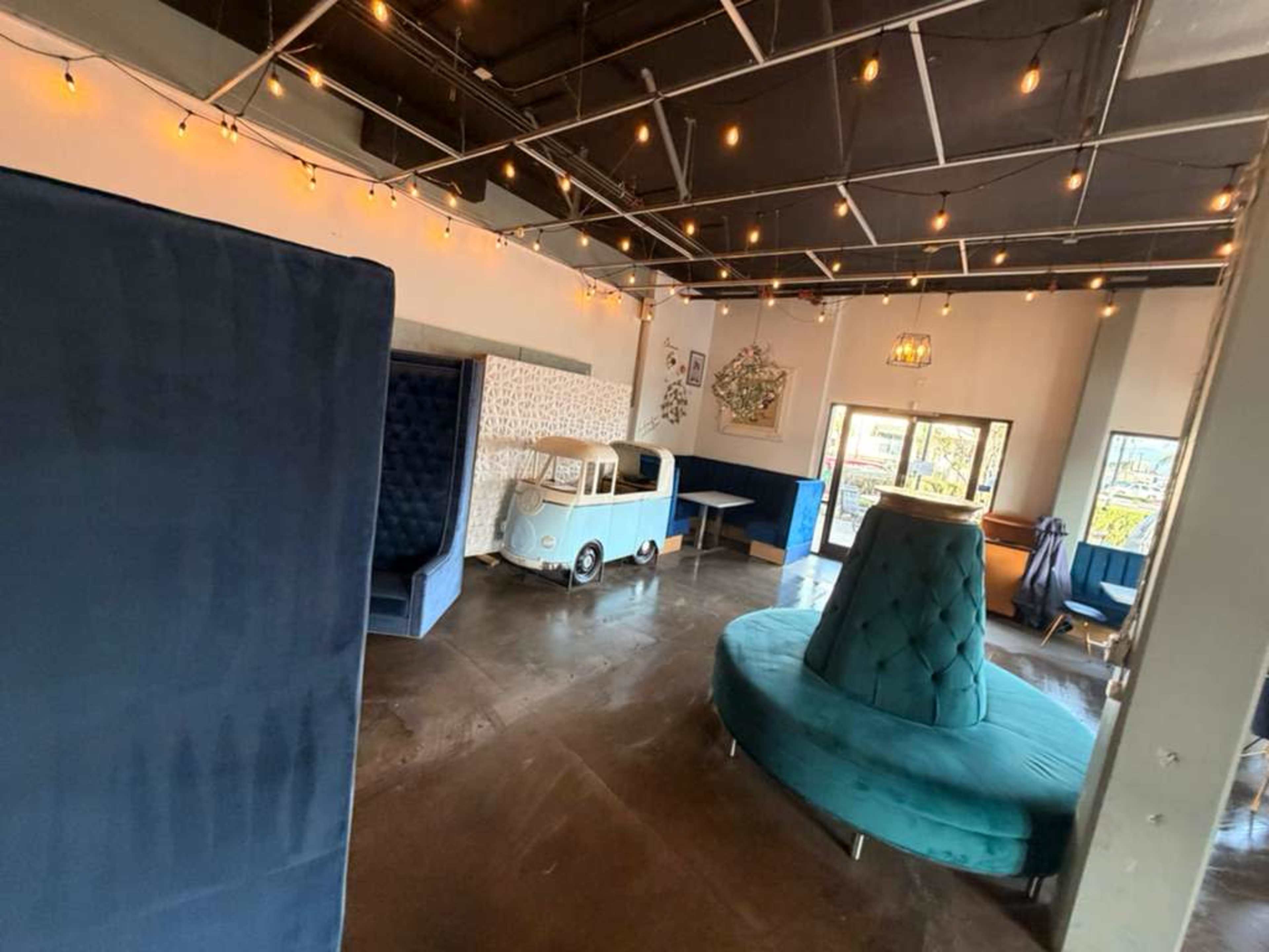 The image shows a modern interior space with plush seating options, including a large circular sofa and private booths, alongside decorative string lights and a vintage cart.