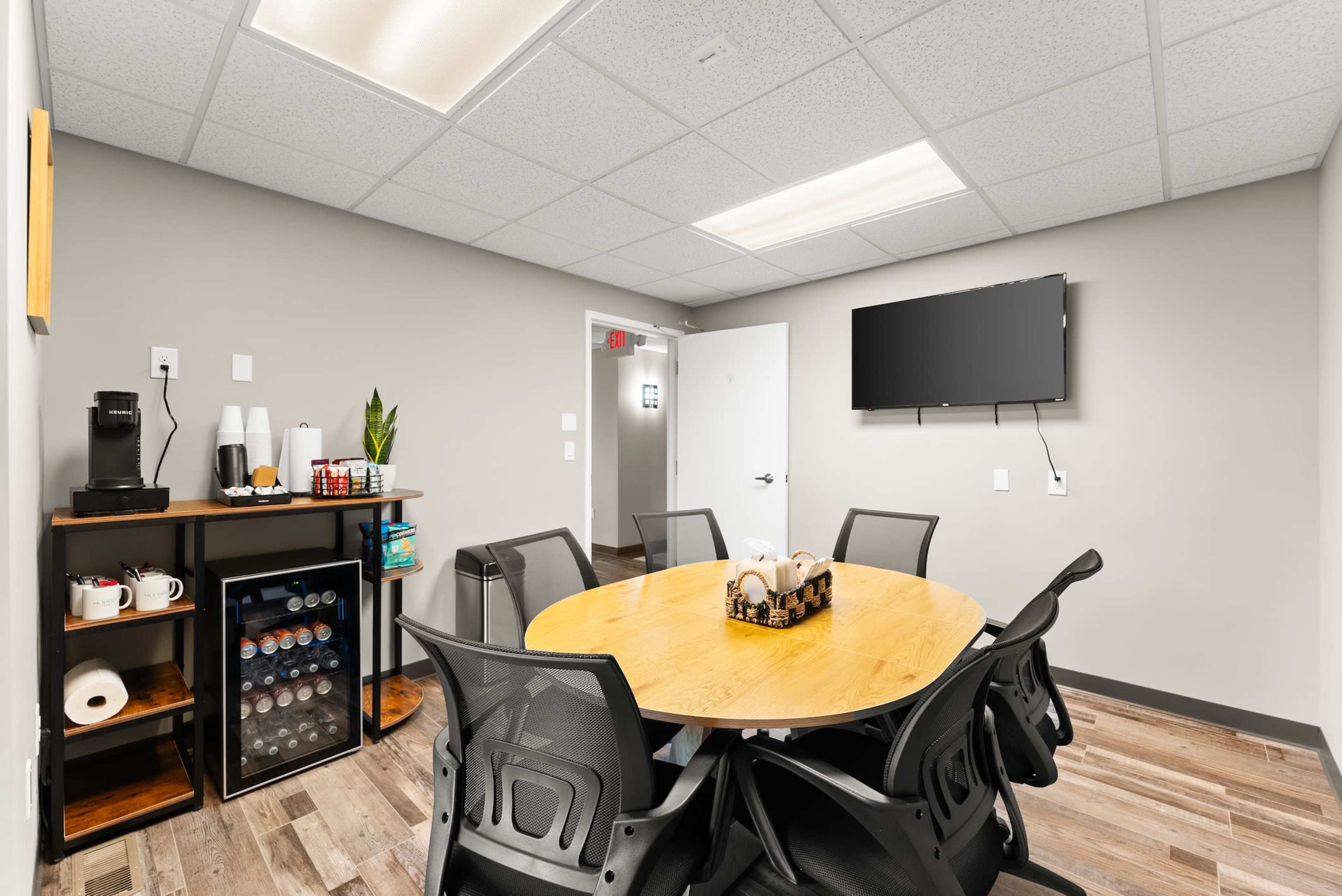 The image shows a modern meeting room with a circular wooden table surrounded by black chairs, a beverage station, and a wall-mounted television.